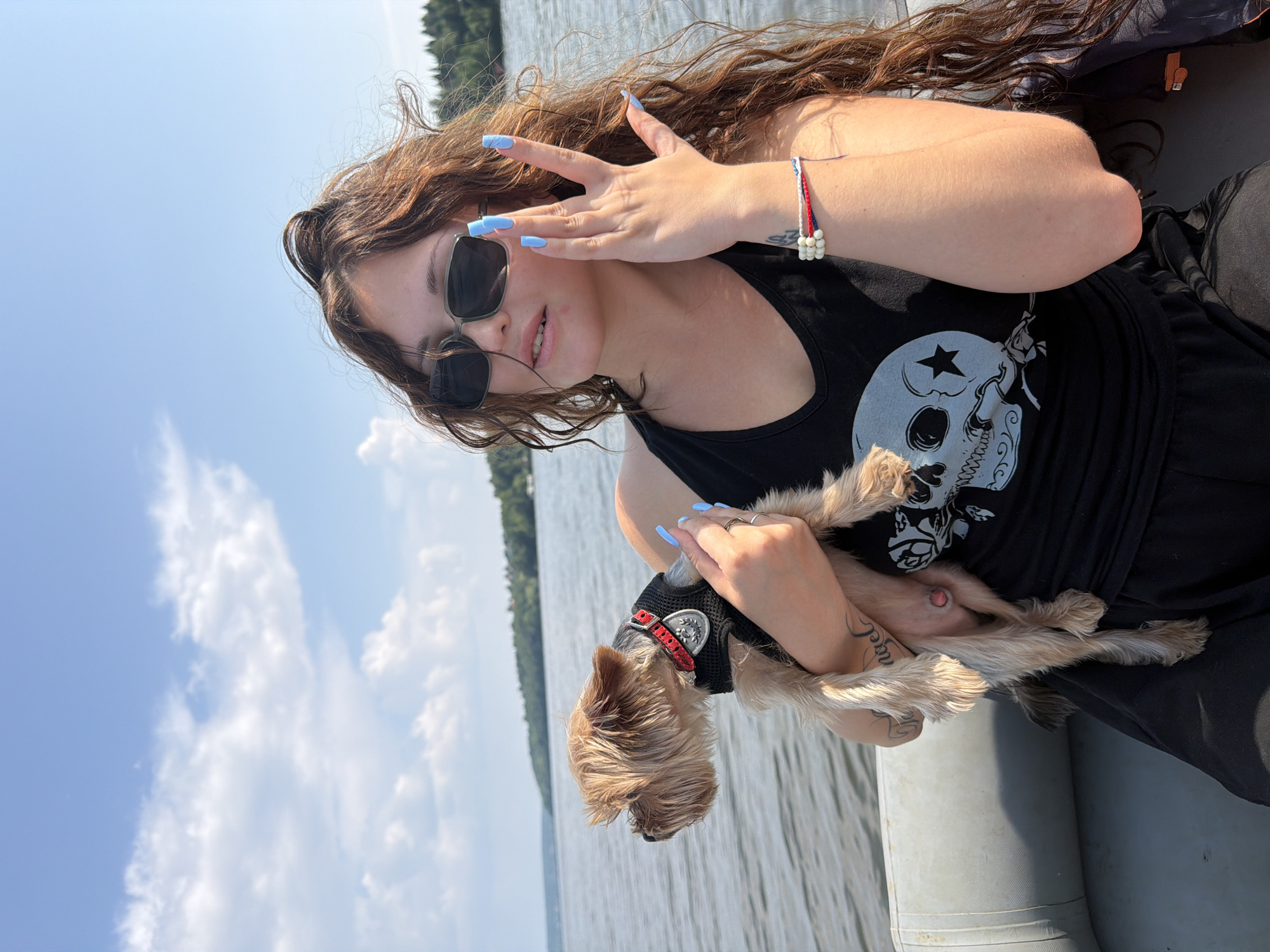 This image, taken in the city of Sylva, Russia, depicts a young woman holding a small dog while on a boat. The woman, with long, wavy brown hair, is wearing a black tank top with a skull graphic, dark sunglasses, and has long, light blue painted fingernails. She is touching her face with her right hand. The dog, a Yorkshire Terrier, is wearing a black harness with a red collar. The boat appears to be an inflatable raft, visible on the left side of the frame. The background shows a body of water, a tree-lined shore, and a blue sky with scattered clouds.