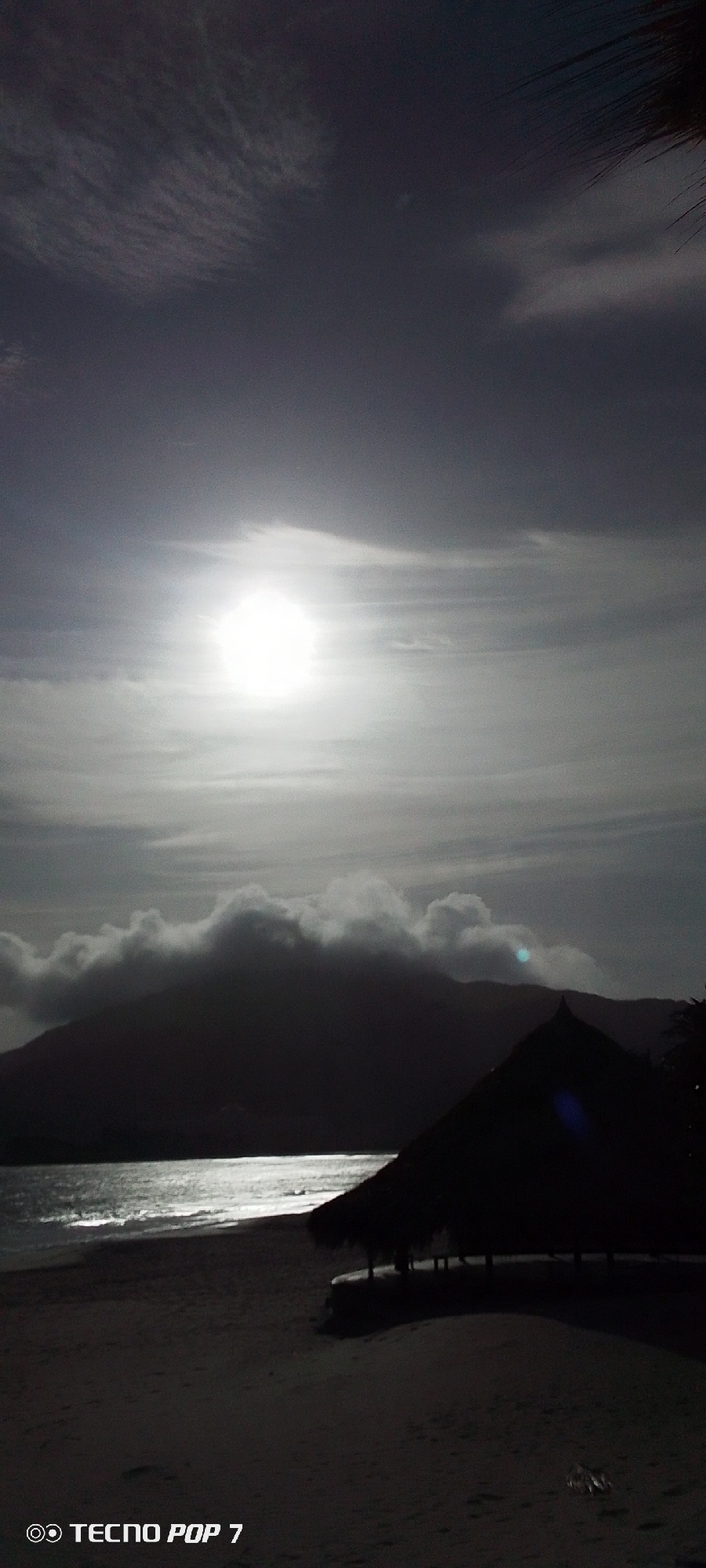 The image shows a beach scene with a bright sun shining through clouds, illuminating the water. In the background is a dark mountain partially covered by clouds. A hut-like structure is located on the beach in the foreground. The image was taken with a TECNO POP 7. It is located in Pedro González, Venezuela.