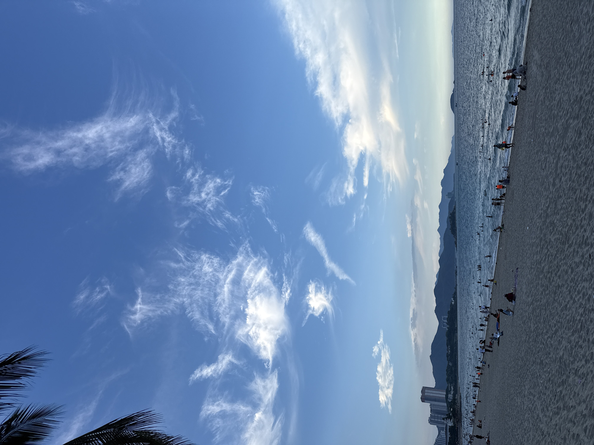 The image depicts a beach scene in Nha Trang Ward, Vietnam, with a clear blue sky filled with wispy white clouds. The ocean stretches to the horizon, meeting a range of hazy, mountainous terrain. The foreground is dominated by a sandy beach where numerous people are visible. Many are standing or walking along the shoreline, while others are wading in the shallow waters of the sea. The water is calm with small waves lapping the shore. The time of day appears to be late afternoon or early evening, judging by the soft light and the long shadows cast by some of the people on the beach. The atmosphere seems relaxed and recreational, with individuals and groups enjoying the seaside. In the distance, a few buildings are visible along the coastline, suggesting a developed urban area nearby. The overall mood is one of a pleasant day at the beach, characterized by natural beauty and human activity.