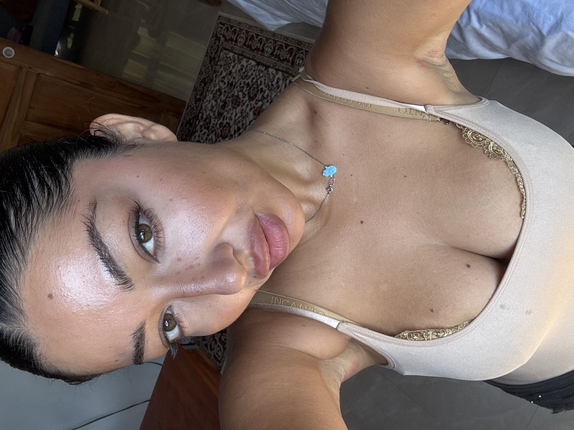 A woman is pictured from an upper angle indoors. She is wearing a beige tank top with lace details around the neckline and a silver necklace with a blue hamsa pendant. She has dark hair pulled back, brown eyes, and full lips. The woman's skin appears dewy. Part of a bed with white sheets is visible in the upper right corner. An intricately patterned rug sits in the middle ground. The floor is tiled. North Kuta, Indonesia is not identifiable in this image.
