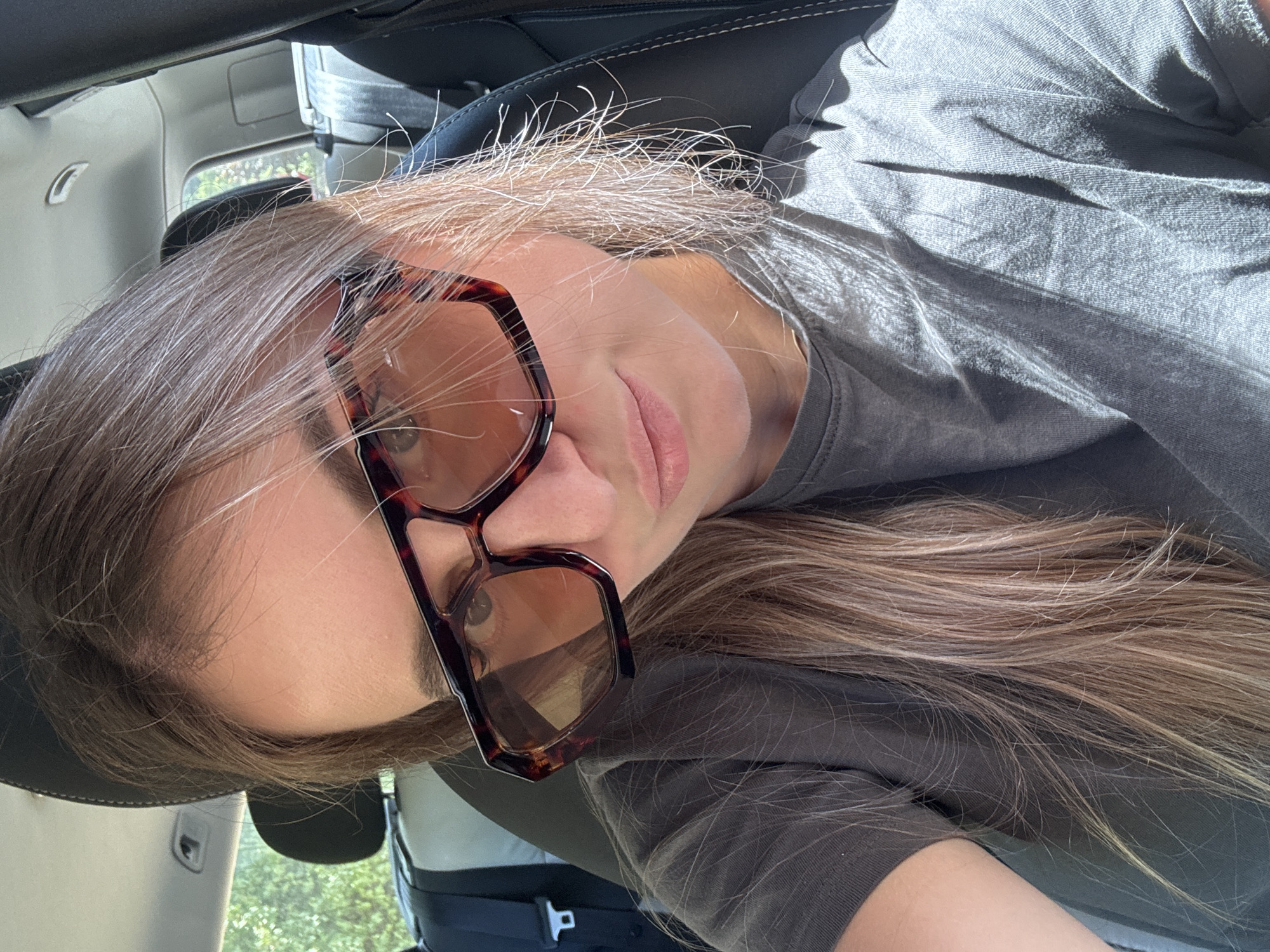 A medium shot photo shows a woman wearing large, tortoise-shell framed sunglasses and a grey t-shirt. Her hair, a mix of brown and blonde highlights, is long and falls over her shoulders. She is inside a vehicle, likely a car, as evidenced by the car seat visible behind her, which is black with white stitching. The background shows the interior of the car, including a headrest and what appears to be part of the car's interior ceiling. Sunlight streams in from the right side, creating highlights on her hair and t-shirt. The image is cropped closely to the woman's face and upper body.