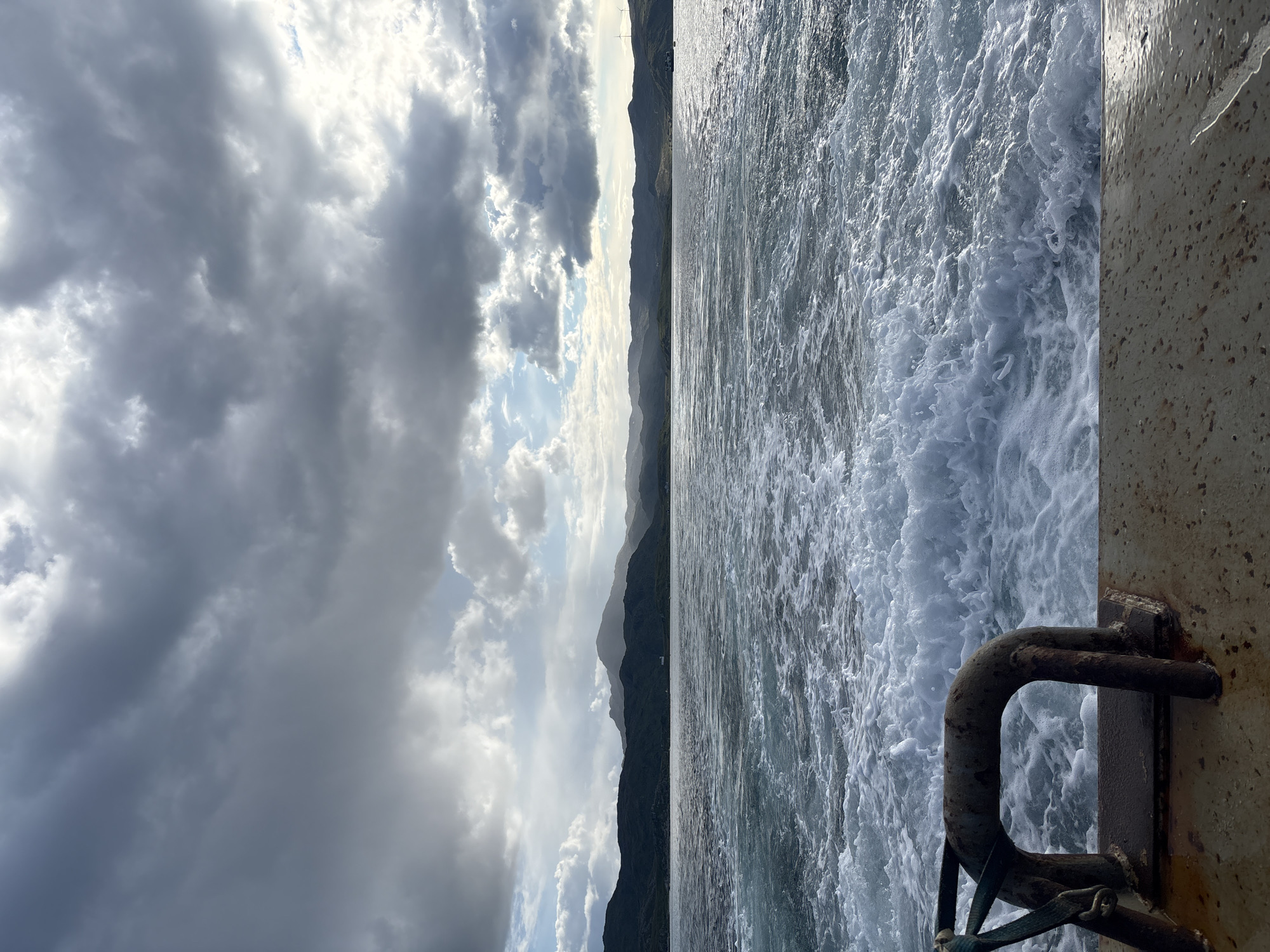 The image depicts a view from the stern of a vessel traveling through choppy, frothy seawater. In the foreground, a weathered, rusty metal cleat with a green rope tied to it is visible on the deck of the vessel. The wake of the boat is prominent, creating white, churning water that extends into the mid-ground. Beyond the water, a range of dark, mountainous islands stretches across the horizon, silhouetted against a dramatic sky filled with heavy, gray clouds interspersed with patches of brighter sky where sunlight breaks through. The overall impression is of a journey through a rugged, coastal landscape under an overcast sky. The location is not specified in the provided context.