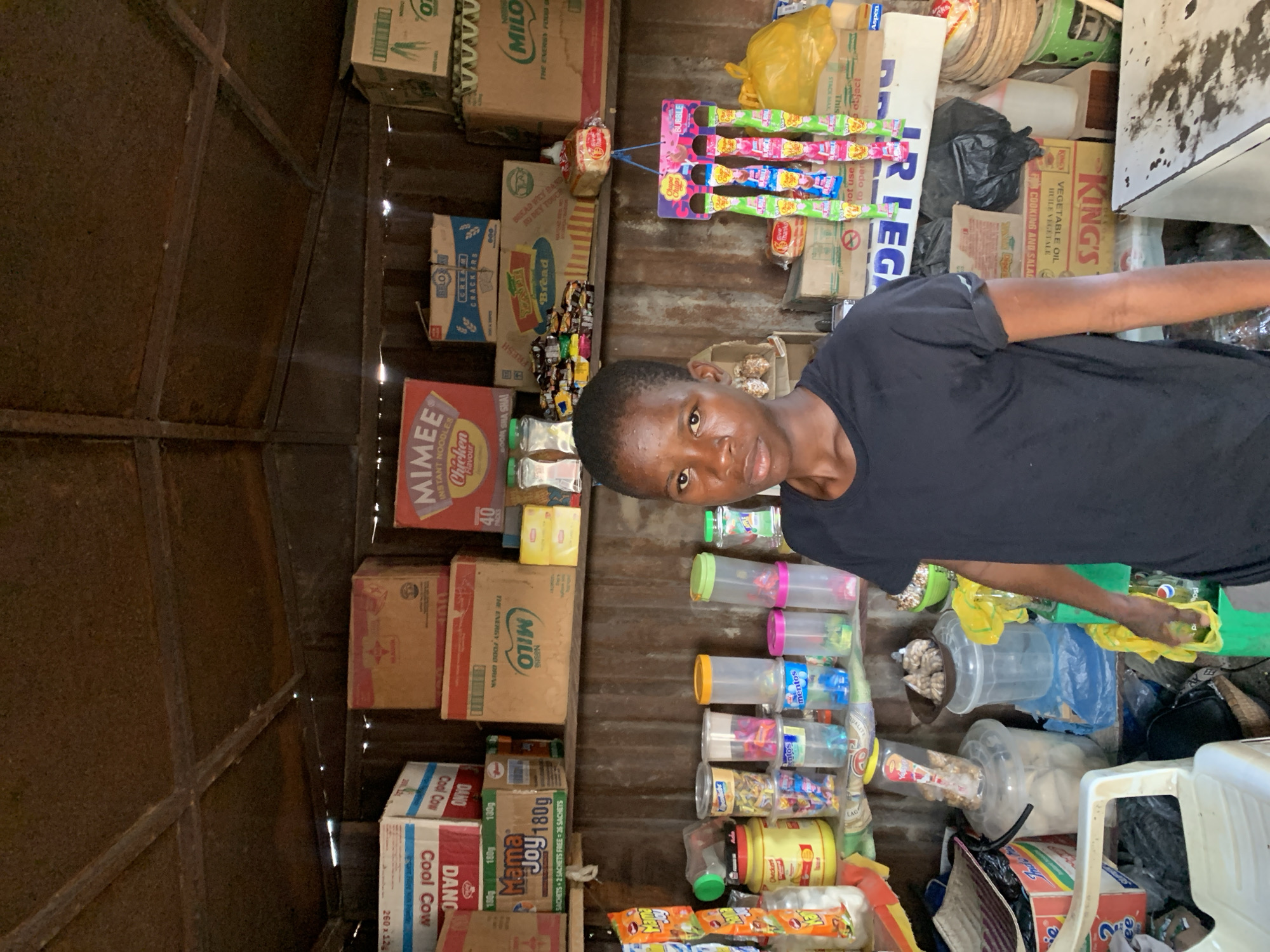 The image depicts a young person in a small shop located in Adankolo, Nigeria. The shop is densely packed with various food items and household goods. Boxes of Milo, Mama Joy, Cool Cow, and various brands of crackers are stacked on shelves. Several containers of candy, snacks, and bubble gum are visible. Other goods include cooking oil and various bags and containers of items. The walls of the shop are made of corrugated metal.