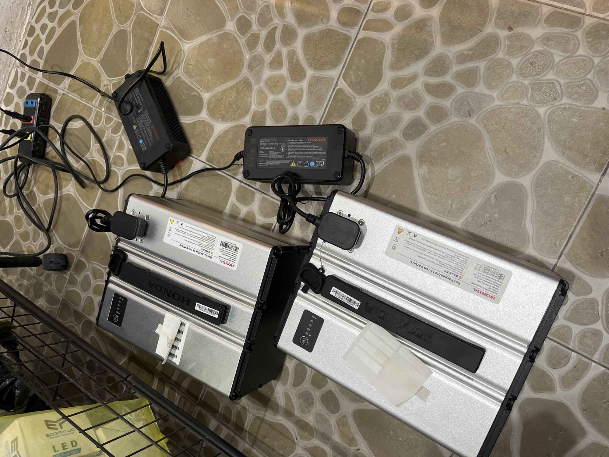 The image shows two Honda rechargeable lithium-ion batteries and their chargers resting on a tiled floor. An extension cord is also visible, with its black cord snaking around the chargers. The batteries and chargers are identified with Honda branding. The background shows a corner with a black metal rack, holding a package labelled "ECOPATUO LED track light New Light Technology". The image was likely taken inside, on a tiled floor. Based on the OCR, one charger has Vietnamese writing. The other battery chargers have English. No people are visible. The precise location within Hội An Ward, Vietnam cannot be confirmed without further information.