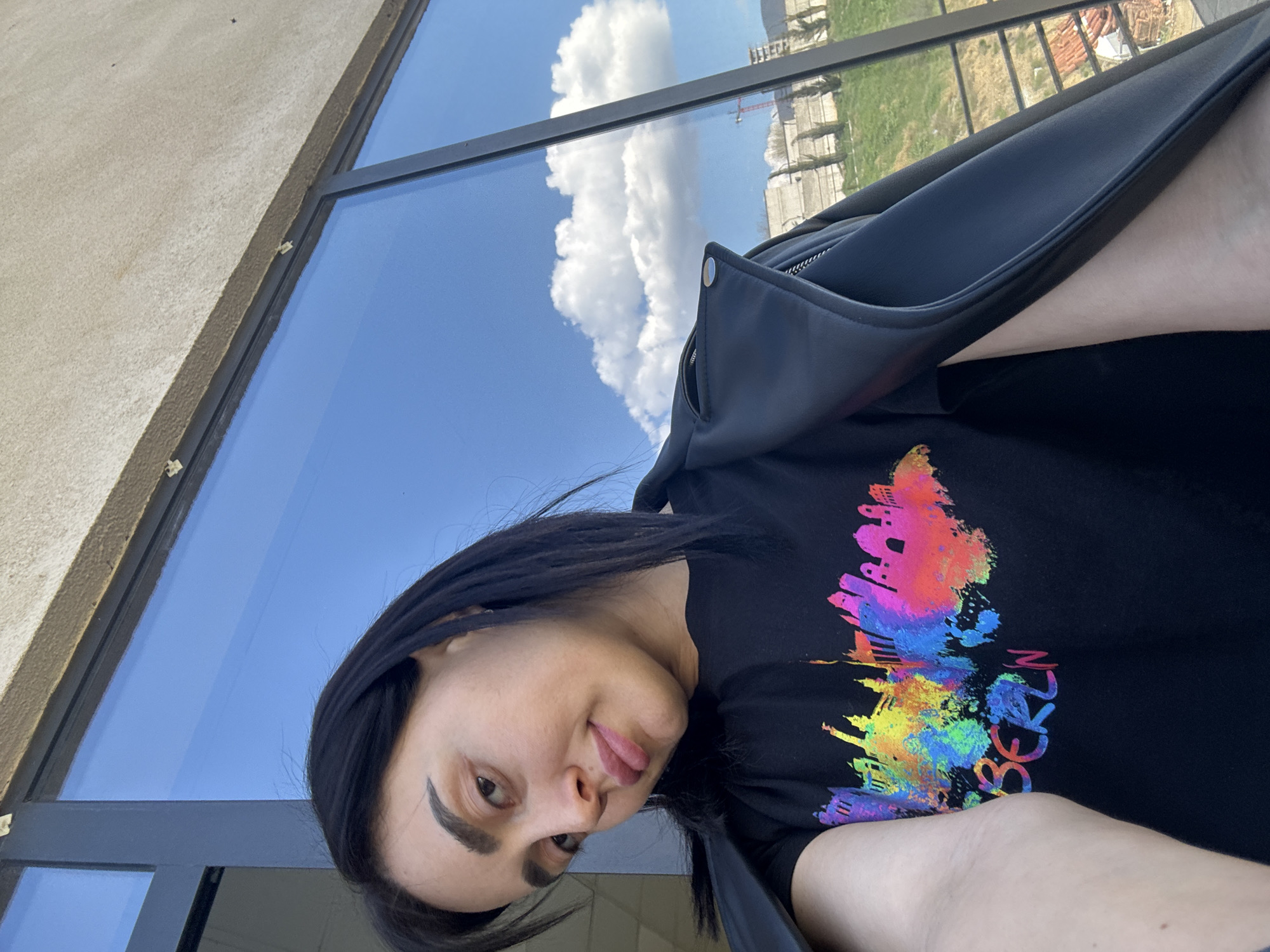 A low-angle shot features a woman with dark hair, wearing a black t-shirt with a colorful, abstract skyline design and a black leather jacket. Her face is partially turned towards the camera with her tongue slightly sticking out. The background shows a clear, blue sky with fluffy white clouds reflected in a large window. Outside the window, a cityscape is visible with buildings, trees, and a crane. The location appears to be Tbilisi, Georgia.