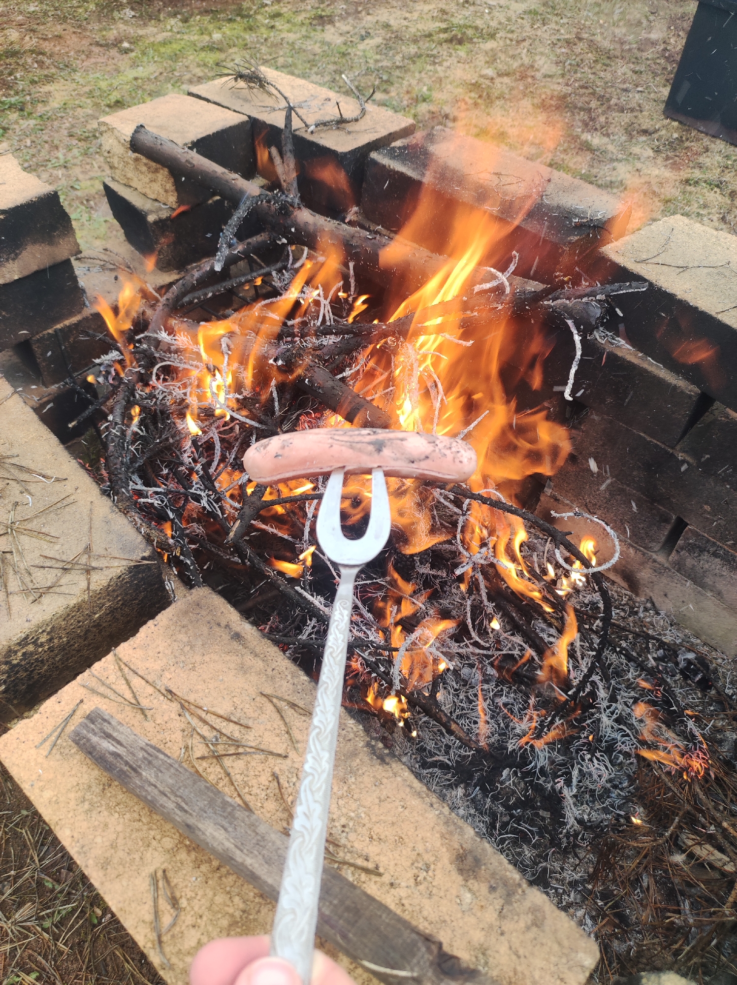 A slightly high-angle outdoor shot focuses on a hot dog impaled on a metal fork, held close to a burning fire. The fire is contained within a makeshift fire pit constructed from light brown bricks. The flames rise intensely, licking at the hot dog and the surrounding twigs and branches that fuel the blaze. The ground surrounding the fire pit shows patches of grass and scattered twigs. In the lower foreground, a hand holds the handle of the fork, suggesting a person is roasting the hot dog. There is no information available that can be used to identify the location as Фоки, Russia.