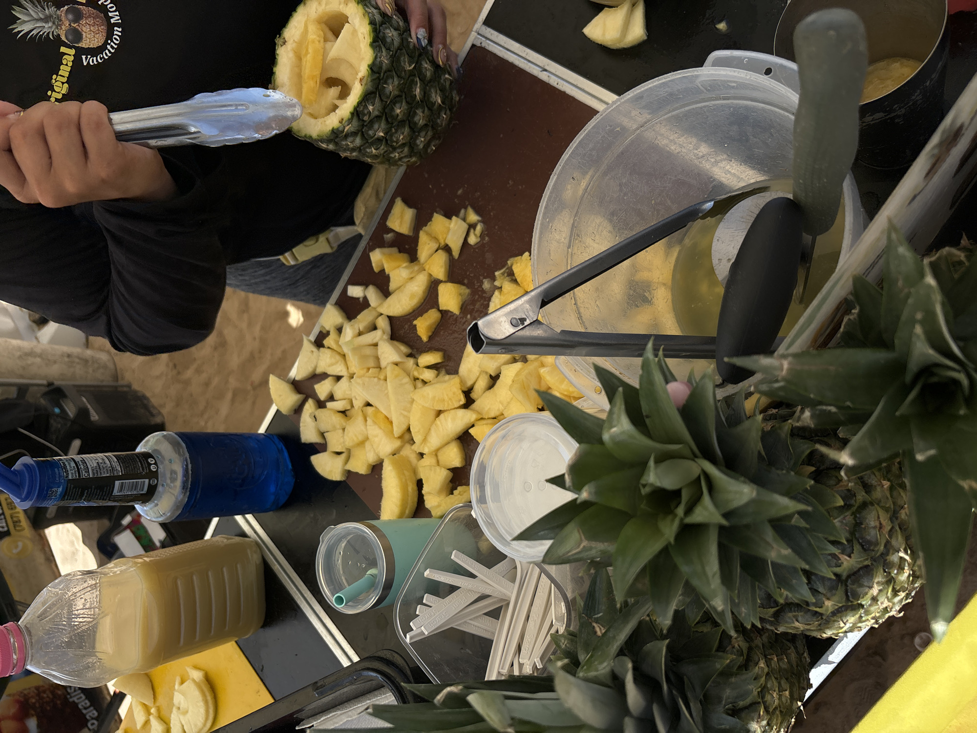 In San Juan, United States of America, a person is preparing pineapple at an outdoor market stall. The scene is set at a food stand, indicated by the presence of various food preparation items and ingredients. In the foreground, several whole pineapples with their distinctive spiky green tops are visible, suggesting they are fresh and ready for processing. Scattered across a dark brown cutting board are numerous pieces of cut pineapple, appearing golden yellow and juicy, ready to be served or used in drinks.
To the left of the cutting board, a large plastic bottle filled with a pale yellow liquid, likely pineapple juice or a beverage, stands upright. Beside it, a smaller blue bottle, possibly containing a cocktail mixer or syrup, is also present. A teal-colored insulated cup with a straw is also on the counter, suggesting a prepared drink. A clear plastic container holds a stack of white stirrers or straws, some labeled with "Plant Starch."
In the mid-ground, a person is using a pair of silver tongs to place pieces of pineapple into a hollowed-out pineapple shell. The person is wearing a black long-sleeved shirt with a graphic featuring a pineapple wearing sunglasses and the text "Original Vacation Mode." Their hands are visible, with painted fingernails.
In the background, some equipment and structures associated with a market stall can be seen, including what appears to be a cart or counter and possibly a canopy or awning. The overall atmosphere suggests a tropical or beachside location, given the abundance of fresh fruit and the casual outdoor setting. The lighting indicates it is daytime.