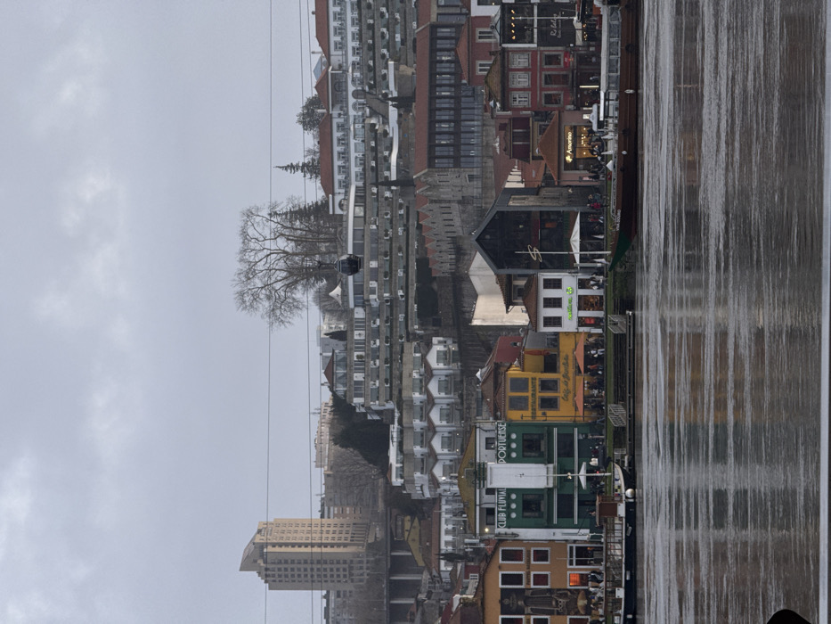 The image depicts a city skyline along a riverbank under an overcast sky. A cable car is visible in the upper left, suspended among bare trees and apartment buildings. Multi-story buildings line the river, including a prominent tall building at the lower left. The river surface reflects the buildings, creating a blurred, mirrored effect. Colorful buildings with awnings are situated at the river’s edge, suggesting a vibrant waterfront area.