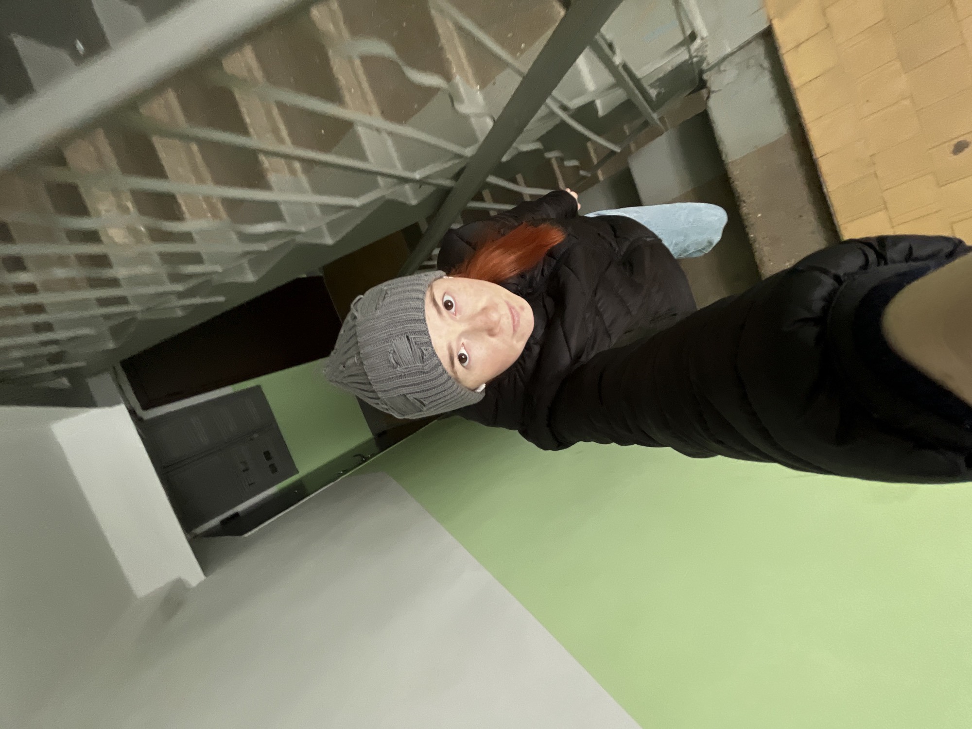 A woman with red hair and a gray beanie is taking a selfie looking upward in what appears to be the stairwell of an apartment building in the city of Chaikovsky, Russia. She is wearing a black, puffy jacket. The stairwell walls are painted a light green and white. The stairs have metal railings. The image includes part of a doorway and a gray electrical panel on the wall. The building's exterior wall is made of tan bricks.