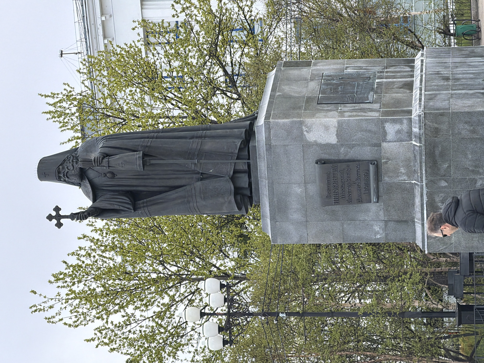 This is a full-shot, eye-level, outdoor photograph of a large bronze statue of a robed man holding a cross in his right hand and a staff in his left. The statue is situated on a grey stone pedestal, which has inscriptions and a bas-relief. Behind the statue, several trees with light green foliage are visible, along with a white building and a street lamp with multiple white globes. In the lower right corner of the image, a man wearing sunglasses and a black puffer jacket is seen from the back, facing away from the statue and towards the right. The statue is located in the city of Magadan, Russia, specifically on the embankment of the Sea of Okhotsk.