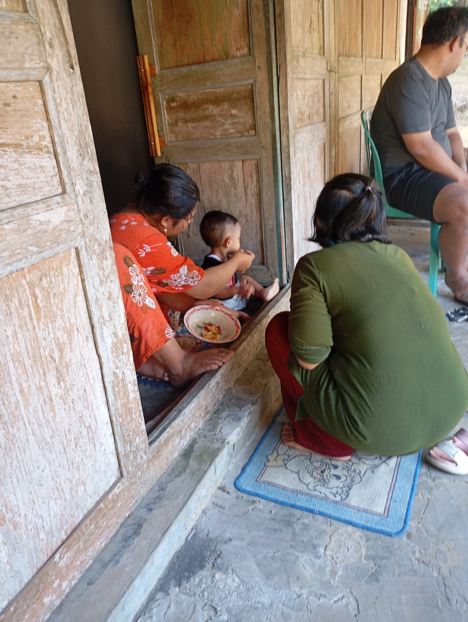 In Ketepung, Indonesia, on a weathered wooden porch, a woman in an orange floral top and pants is feeding a baby. The baby, wearing a striped shirt, sits facing away from the camera. Beside the woman is a small bowl containing food. Another woman, wearing a green t-shirt and red pants, kneels on a blue mat featuring a cartoon character. To the right, a man in a dark t-shirt and shorts sits on a green plastic chair. The environment is rustic, with wooden doors and a concrete floor, suggesting a simple dwelling.