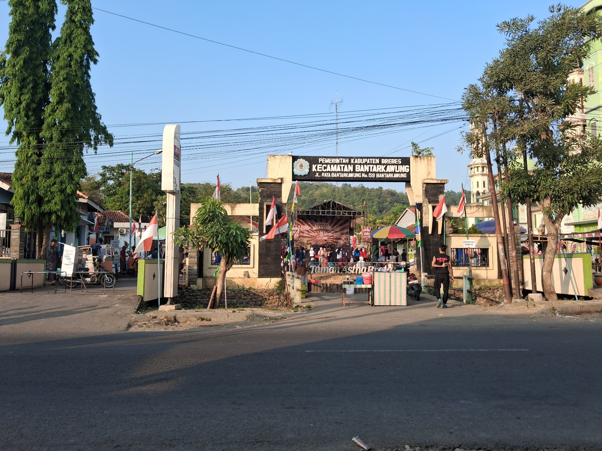 The image shows an archway entrance to an area labeled "PEMERINTAH KABUPATEN BREBES KECAMATAN BANTARKAWUNG" in Bantarkawung, Indonesia. The archway has flags and banners, including Indonesian flags. Beneath the arch, there is a stage with people gathered. To the left of the arch is a tall white signpost. To the right, there are trees and a green building with a minaret, suggesting a mosque or religious structure. In the foreground, there is a road with sunlight casting shadows. A few people are visible walking and standing near stalls set up in front of the archway. One man in a black shirt with red lettering that reads "RUNNING CLUB" is walking away from the camera. A motorcycle is parked on the left side. The environment appears to be a street scene in a town or city.