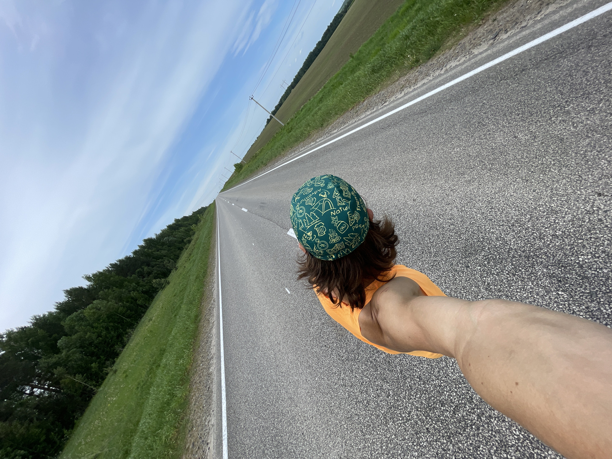 The image shows a person standing on a paved road in Перевозное, Russia. The person is wearing a green hat with camping-related designs and an orange shirt. The road stretches out towards the horizon, with green fields and trees on either side. Power lines are visible along the roadside. The sky is blue with some clouds.