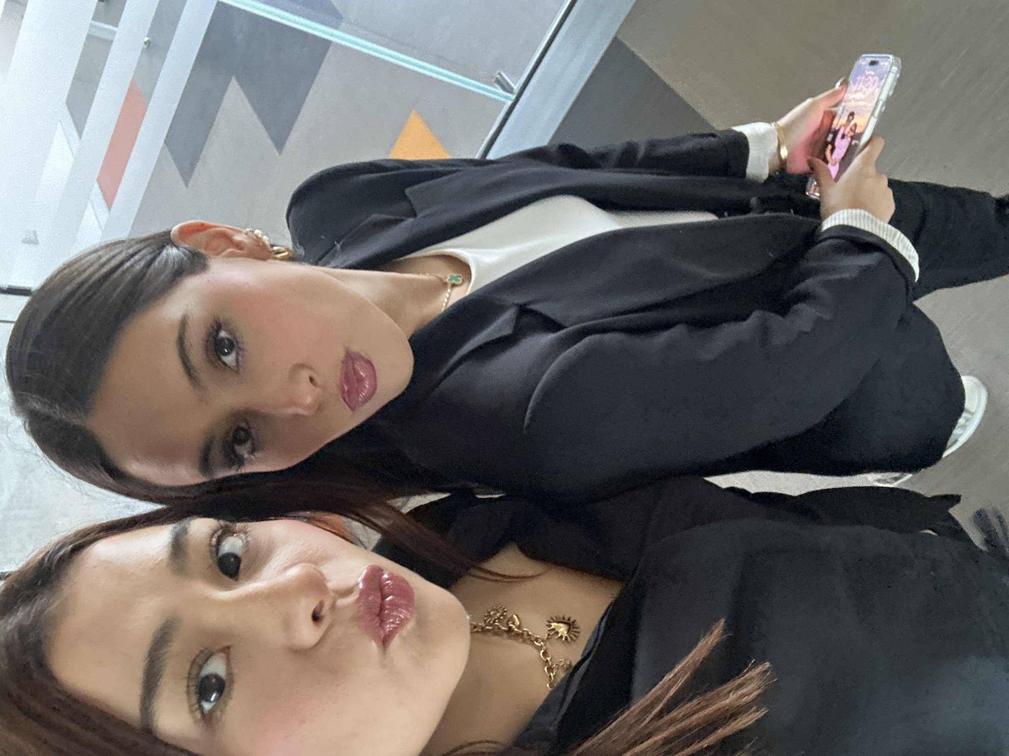 Two women are posing for a selfie indoors. The top woman has dark brown hair pulled back, and is wearing a black blazer over a white shirt, with gold earrings and a delicate necklace. She is holding a phone with a time displayed on the screen and wearing bracelets. The bottom woman has shoulder-length brown hair and is also wearing a black blazer with a gold necklace. The background appears to be a modern interior, potentially an office building, with a glass window or panel. The location may be San Bernardino Tlaxcalancingo, Mexico.