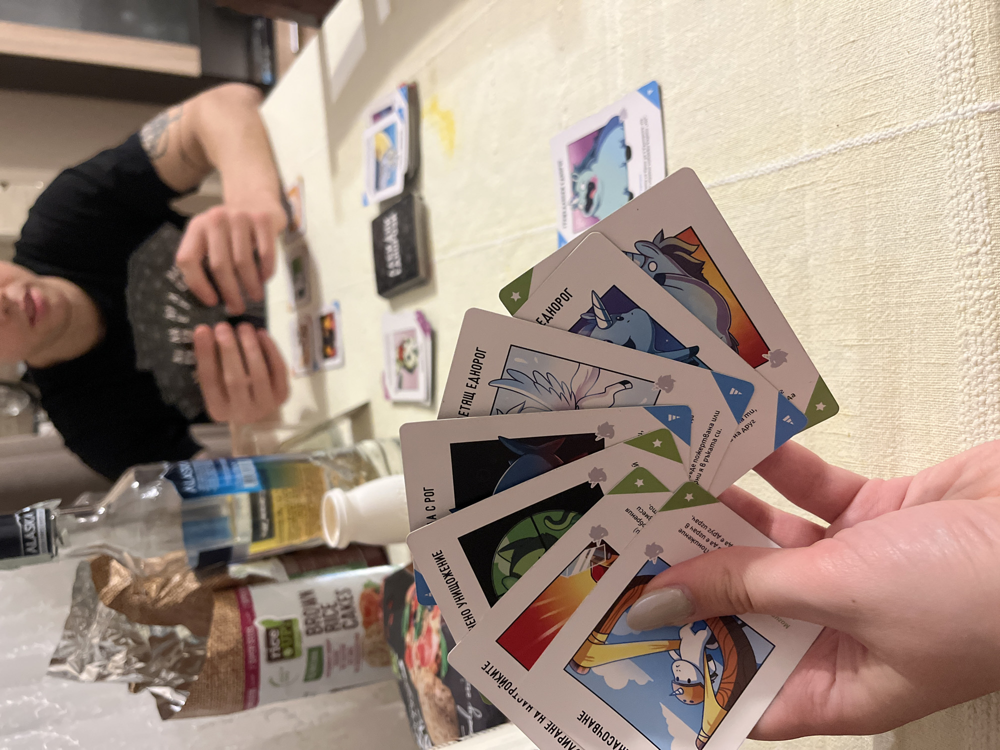 A tabletop scene features a person playing a card game. A hand holds a fan of playing cards depicting unicorns. The cards contain text in Cyrillic, specifically referencing "единорог" (unicorn). Other cards are scattered on the textured, off-white tabletop. Two individuals appear to be engaged in the game. Additional objects on the table include an open box of brown rice cakes and two empty liquor bottles bearing the name "ALASKA." The setting is residential, possibly Varna, Bulgaria.