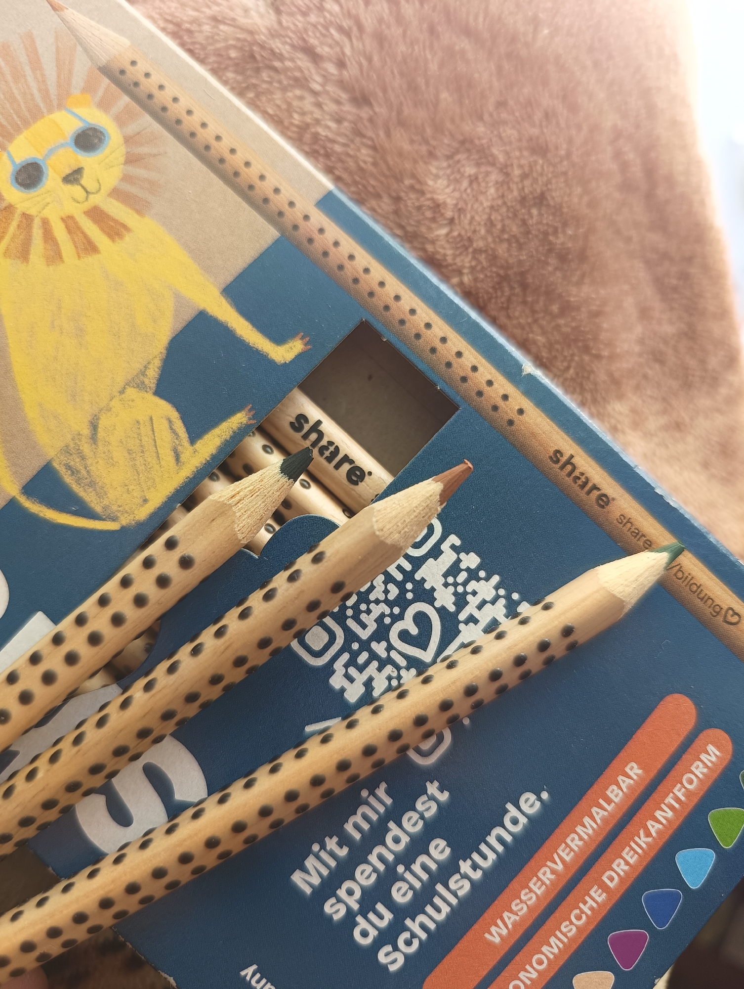 The image presents a close-up view of a blue box containing colored pencils, resting on a soft, light brown, textured fabric surface. The box lid features a cheerful, stylized yellow lion with a prominent mane and blue sunglasses. Several wooden pencils, each distinguished by a pattern of small black dots along its natural wood body, are visible. Some pencils are sharpened, showing black, brown, and green tips.
The brand name "share" is visible on the pencils and repeatedly printed on the box, often accompanied by "share/bildung" (share/education) and a heart icon. German text on the blue part of the box reads, "Mit mir spendest du eine Schulstunde." (With me, you donate a school lesson.), indicating a social contribution. Further text highlights product features: "WASSERVERMALBAR" (water-soluble, suggesting watercolor pencils) and "ONOMISCHE DREIKANTFORM" (ergonomic triangular shape). A QR code is also clearly visible on the box.
The scene captures a new product, well-lit with natural light, emphasizing its unique design, functional features, and philanthropic mission. There is no specific activity taking place; it is a still life photograph of the product packaging and its contents. The overall impression is one of a creatively designed, functional product with a positive social message.