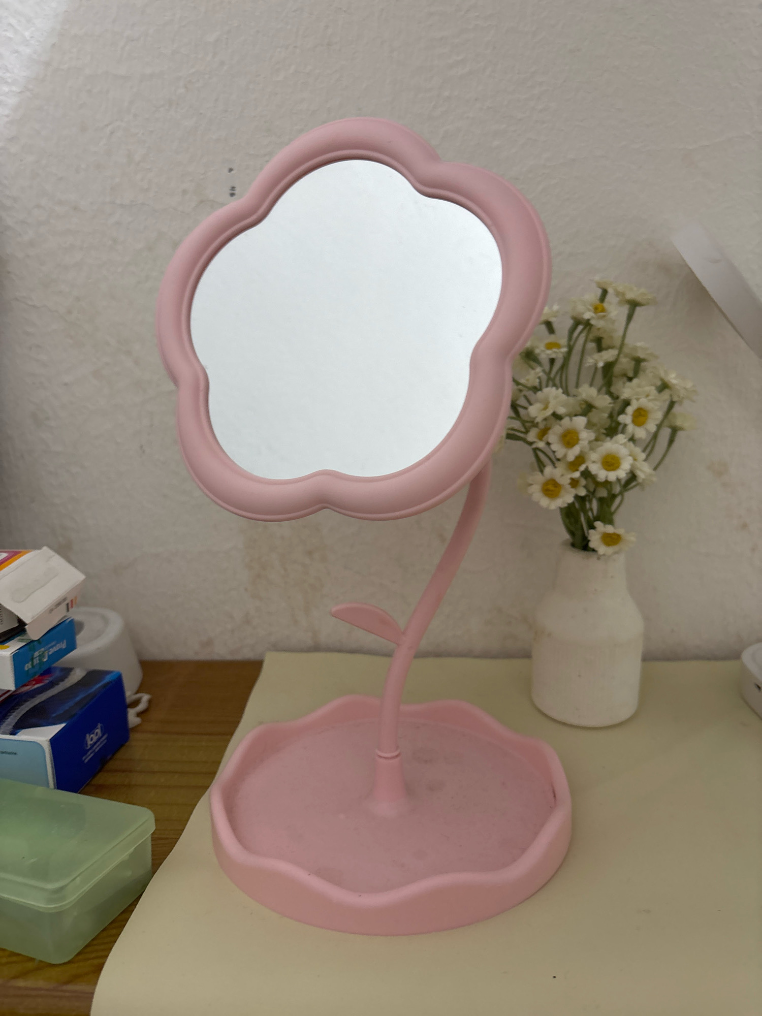An indoor medium shot captures a pink flower-shaped makeup mirror on a desk. The mirror has a scalloped edge and stands on a flexible stem attached to a shallow, flower-shaped base. To the right of the mirror, a white vase holds a bouquet of small white daisies. In the background, a textured wall shows a few dark marks. On the desk to the left, a stack of small boxes is partially visible, with a green plastic container in front of them. The desk surface is a pale yellow, and the wall behind it is off-white. The scene is located in Wanajaya, Indonesia.