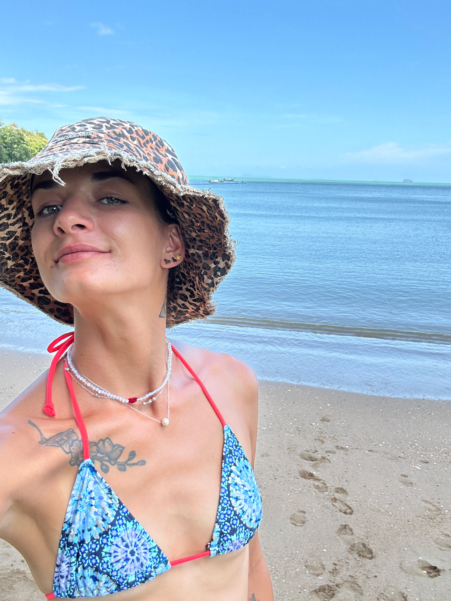 A woman with short dark hair, wearing a leopard-print bucket hat and a blue patterned bikini with red trim, stands on a sandy beach in Ratsada, Thailand. She has several necklaces on and a tattoo on her left shoulder. The ocean is visible behind her, with a clear blue sky overhead. Footprints are visible in the sand.