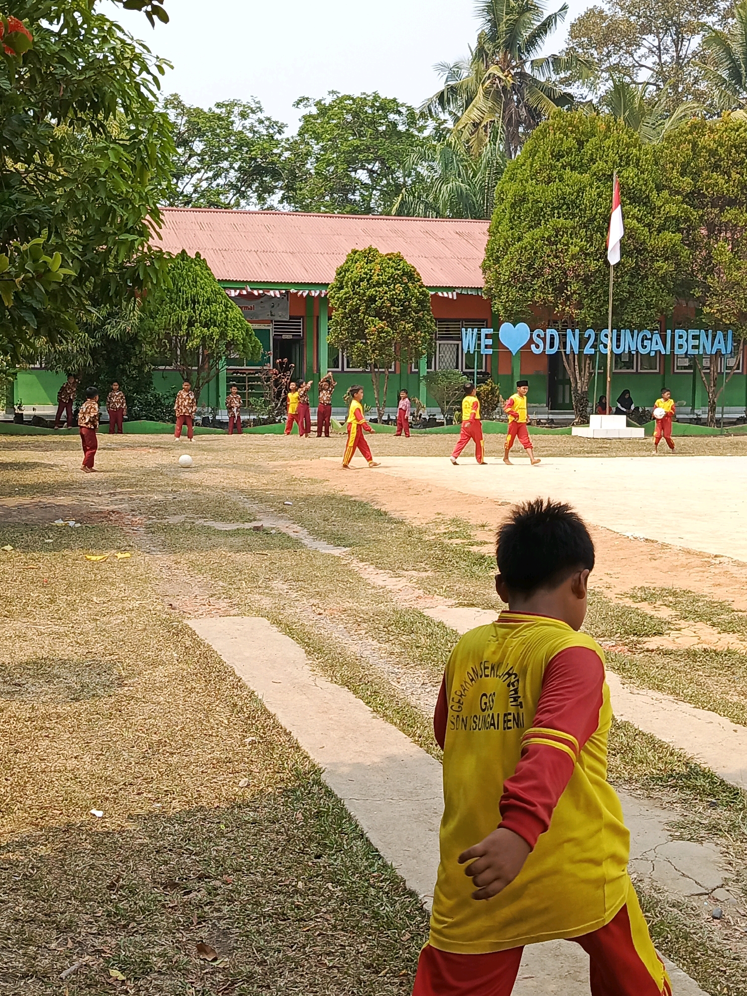 The image depicts an outdoor scene at an elementary school, likely SD Negeri 2 Sungai Benai, in Megang Sakti IV, Indonesia. In the foreground, a young boy with dark hair, wearing a yellow long-sleeved shirt with red sleeves and red pants, is walking away from the camera. The back of his shirt has white text. Several other children, appearing to be students, are scattered across a field in the midground. Some are wearing similar uniforms of brown and red patterned shirts and red pants, while others are in yellow and red athletic wear. A white soccer ball is visible on the dry, grassy field, suggesting a game or practice is in progress. In the background, a long, single-story building with a reddish-orange roof and green trim serves as the schoolhouse. A flagpole with an Indonesian flag is positioned in front of the school. A large blue sign with white text and a blue heart shape reads "WE ♥ SD N 2 SUNGAI BENAI". Lush green trees and palm trees surround the school building and the field, indicating a tropical or subtropical environment. The sky is bright and clear, suggesting a sunny day. The overall scene conveys a sense of a school day with children engaged in outdoor activities.