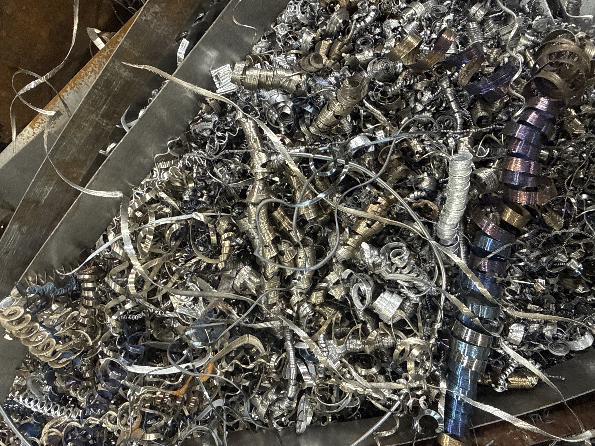 The image depicts a large quantity of metal shavings and turnings inside a metal container. The shavings vary in color from silver to gold to iridescent blues and purples, indicating different types of metal or heat treatment. The shapes are diverse, including tight spirals, loose coils, and thin, ribbon-like strands. The container appears to be partially full, suggesting it is a receptacle for metal waste from machining processes. There is no indication this is in Aramil, Russia.