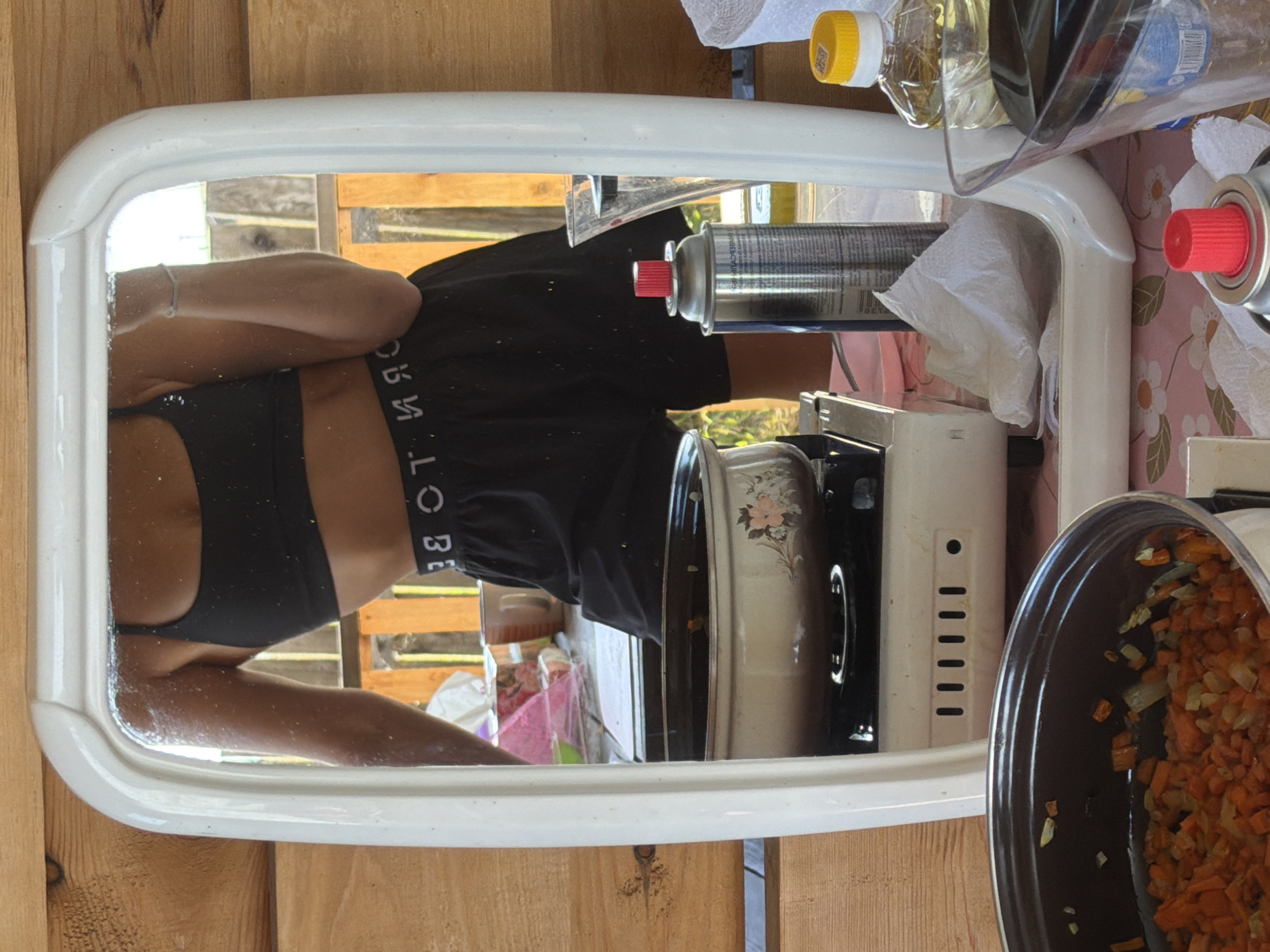 This is a vertical, close-up, medium shot of a person's torso reflected in a white-framed mirror. The subject is wearing a black sports bra and black shorts. The waistband of the shorts has white text that reads "CORE". The person's arm is visible, resting on a surface.
The setting appears to be outdoors or in a rustic indoor space, with wooden planks visible behind the mirror. In the mirror's reflection, other objects are visible on a table or counter, including a clear plastic bottle with a yellow cap, a spray can with a red nozzle, and what appears to be a portable stove. A pot containing chopped carrots and onions is also reflected. The overall lighting suggests it is daytime, and the weather is not discernible. There are no explicit emotional cues visible from the cropped image.