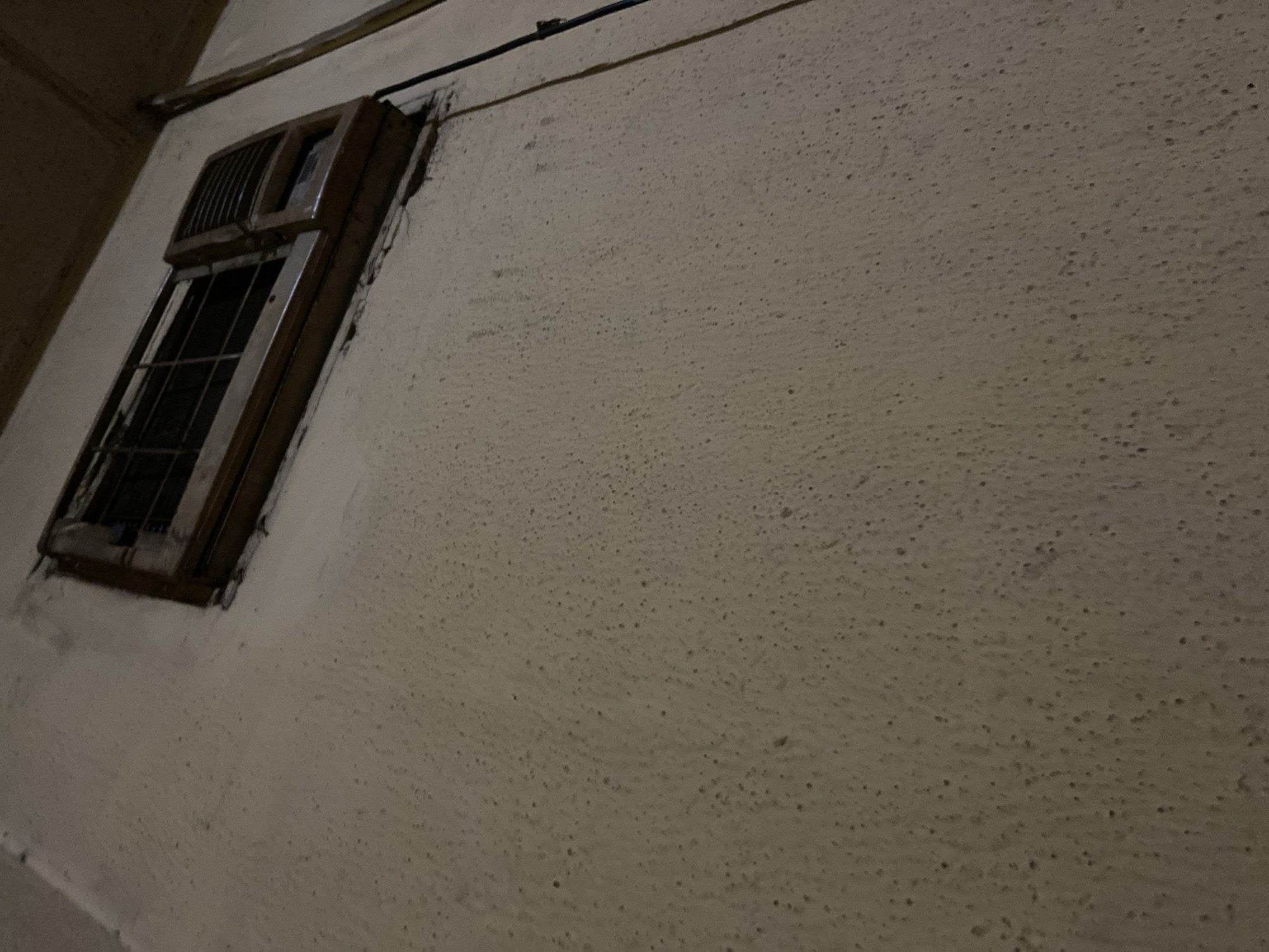 The image shows an outdoor scene at night, likely in Jalingo, Nigeria. A rough, textured, cream-colored wall occupies most of the frame. Embedded in the upper right portion of the wall is a window air conditioning unit, appearing to be made of wood and plastic. The unit has a metal grate on the left side and louvers on the right. A black electrical cord hangs down from the unit, running vertically along the wall. The wall itself is textured with numerous small, dark pores or irregularities. The lighting is dim, suggesting it is nighttime, and the overall atmosphere is somewhat grimy.