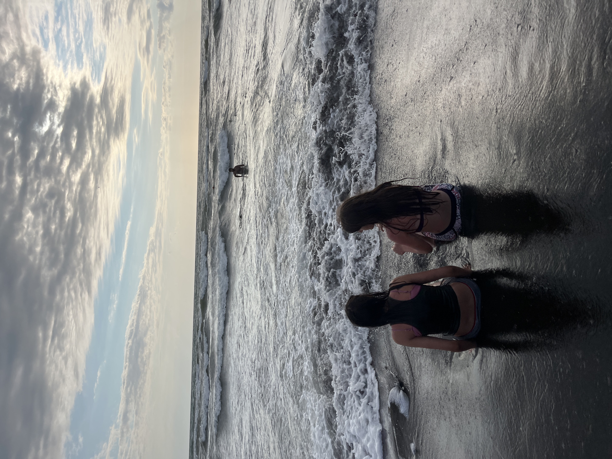 This media file shows a beach scene with two young girls sitting in shallow water. The girls are facing away from the camera, and their backs are to the viewer. One girl is wearing a dark tank top with a pink bikini top underneath and patterned shorts, while the other is wearing a black bikini top and patterned bottoms. They are sitting in the water at the edge of the ocean, with waves gently lapping around them.
In the background, a lone figure can be seen wading in the ocean. The water is a dark, shimmering grey, reflecting the overcast sky. The sky is filled with dramatic, grey clouds, with patches of lighter sky peeking through, suggesting either early morning or late afternoon light. The overall mood is peaceful and serene, with the sound of the ocean implied by the gentle waves.
The location is Ureki, Georgia, known for its magnetic black sand beaches. The sand in the foreground appears dark and wet, consistent with this location. There is no visible text in the media file. The activity is simply enjoying the water at the beach.