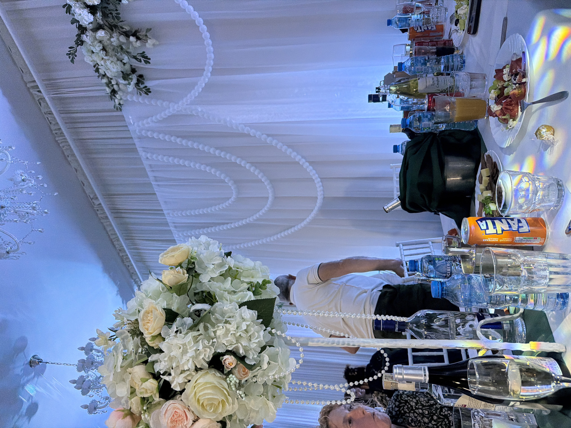 The image depicts a banquet-style table setting, likely within a wedding or celebratory event, located in Plovdiv, Bulgaria. The table is covered with a reflective surface, possibly mirrored or highly polished. Arranged on the table are multiple bottles of water and other beverages, including wine and juice. Glasses of various shapes and sizes are present. Plates with food, including a salad with cured meat and olives, are also on display.
Decorative elements are prominent, including a large floral arrangement in the foreground with white and peach-colored roses. Strands of white faux pearls are draped across the table and around the floral display. Above the table, a white fabric canopy is decorated with more flowers and strands of pearls.
Two people are partially visible. A man in a white shirt is leaning over the table, possibly adjusting or arranging items. A woman with blonde hair, wearing a dark patterned top, is visible at the bottom of the frame, possibly seated at the table.
The lighting appears to be artificial, with strong reflections and shadows suggesting a venue or event hall. A chandelier is visible in the background.