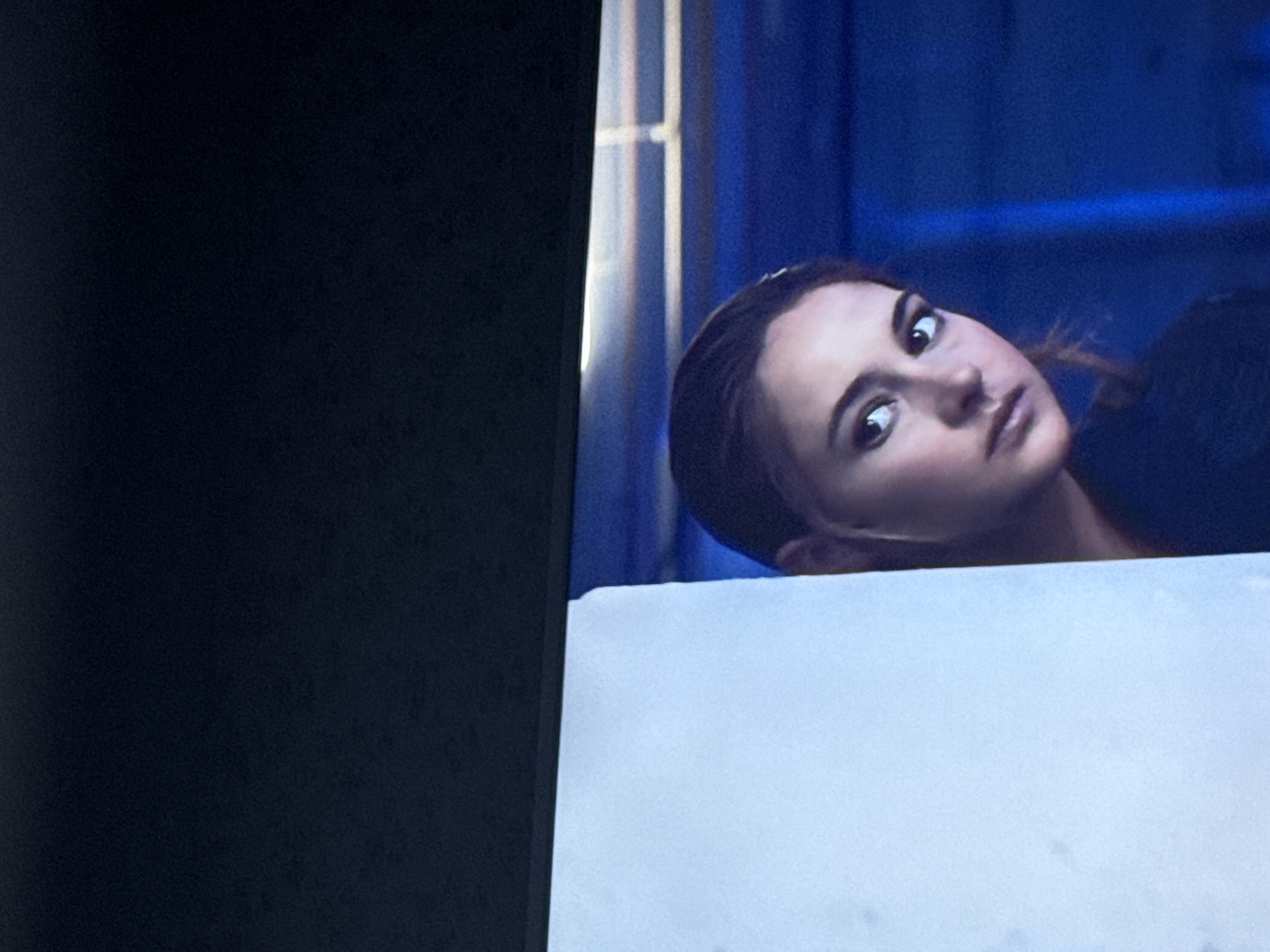 The image shows a television screen displaying a scene with a woman with brown hair and light skin looking at the camera. The background features blue curtains and a white structure is in the foreground. There is nothing to indicate this image was taken in Mytishchi, Russia, or any other geographic location.