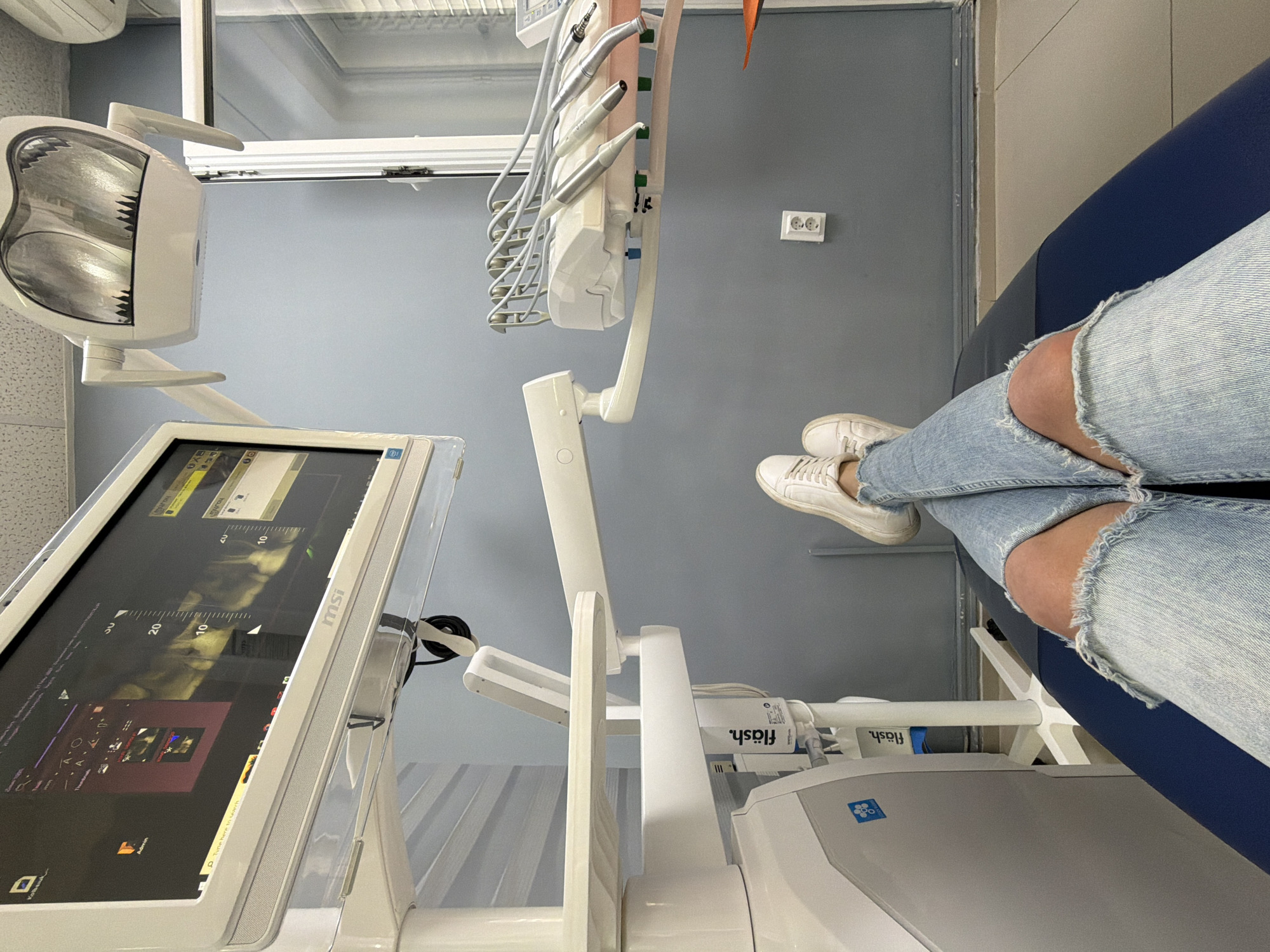 A first-person, overhead perspective presents a person seated in what appears to be a dentist's chair. The individual is wearing ripped light-blue jeans and white sneakers. The dental equipment surrounds the person, featuring a monitor labeled "msi", a dental light, and various dental instruments. A device labeled "fläsh" is also visible. The room has a neutral, sterile color scheme with grey walls. The location is most likely a dentist's office, though the city of Budva, Montenegro is not evident in the image.