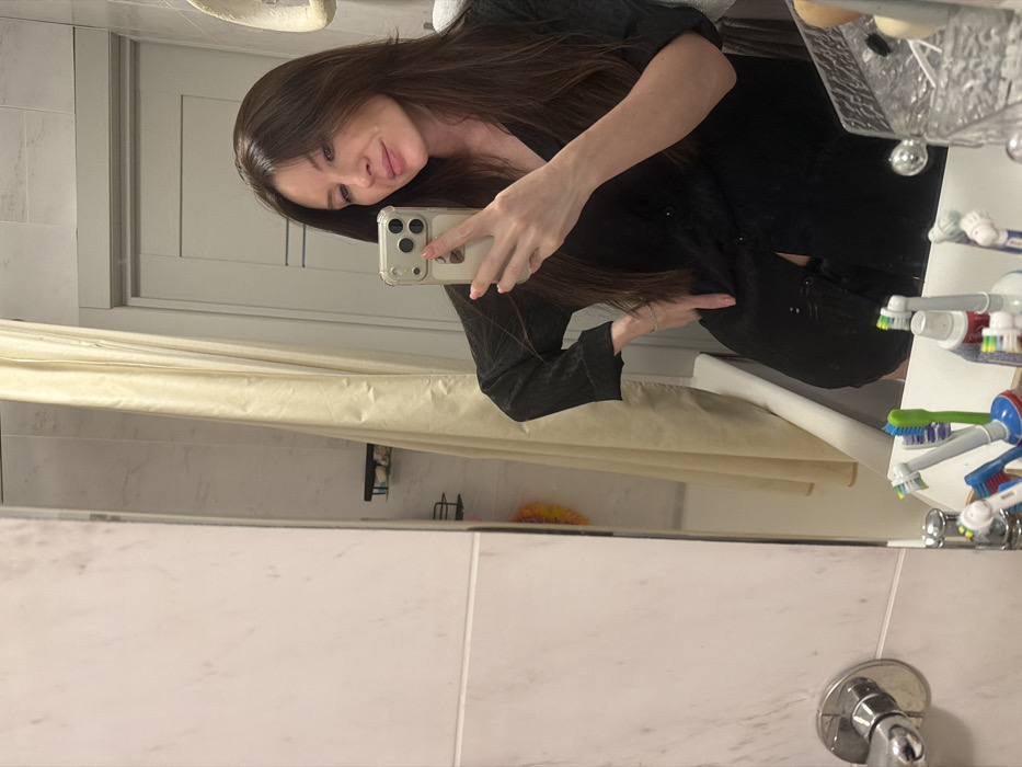 A woman in a dark top takes a mirror selfie in a bathroom. She holds her phone with her right hand, posing slightly. The bathroom features white tile walls, a shower curtain, and a countertop with toothbrushes and other toiletries visible. The overall scene is an everyday moment captured through the reflection.