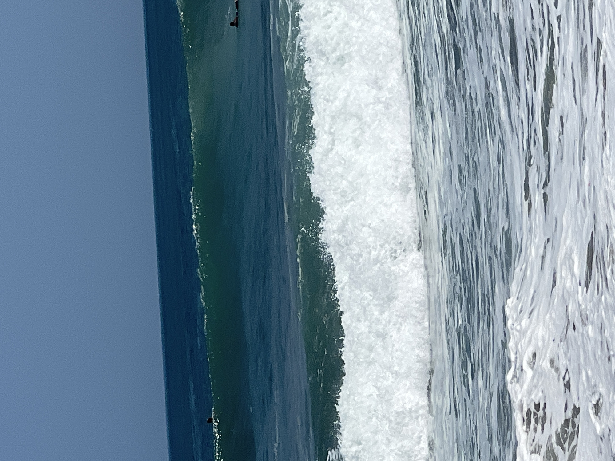 In Kuta, Indonesia, a wide shot captures the vast expanse of the ocean on a clear day, with the horizon dividing the deep blue sky from the dark blue sea. In the middle ground, a large, dark blue wave is cresting, its translucent green face illuminated by the sun. Further out, a surfer is visible, paddling on their board. Closer to the shore, a smaller, foamy white wave is breaking, creating a frothy white wash that extends towards the foreground. The water in the foreground is a lighter blue, with ripples and small whitecaps indicating the movement of the waves.