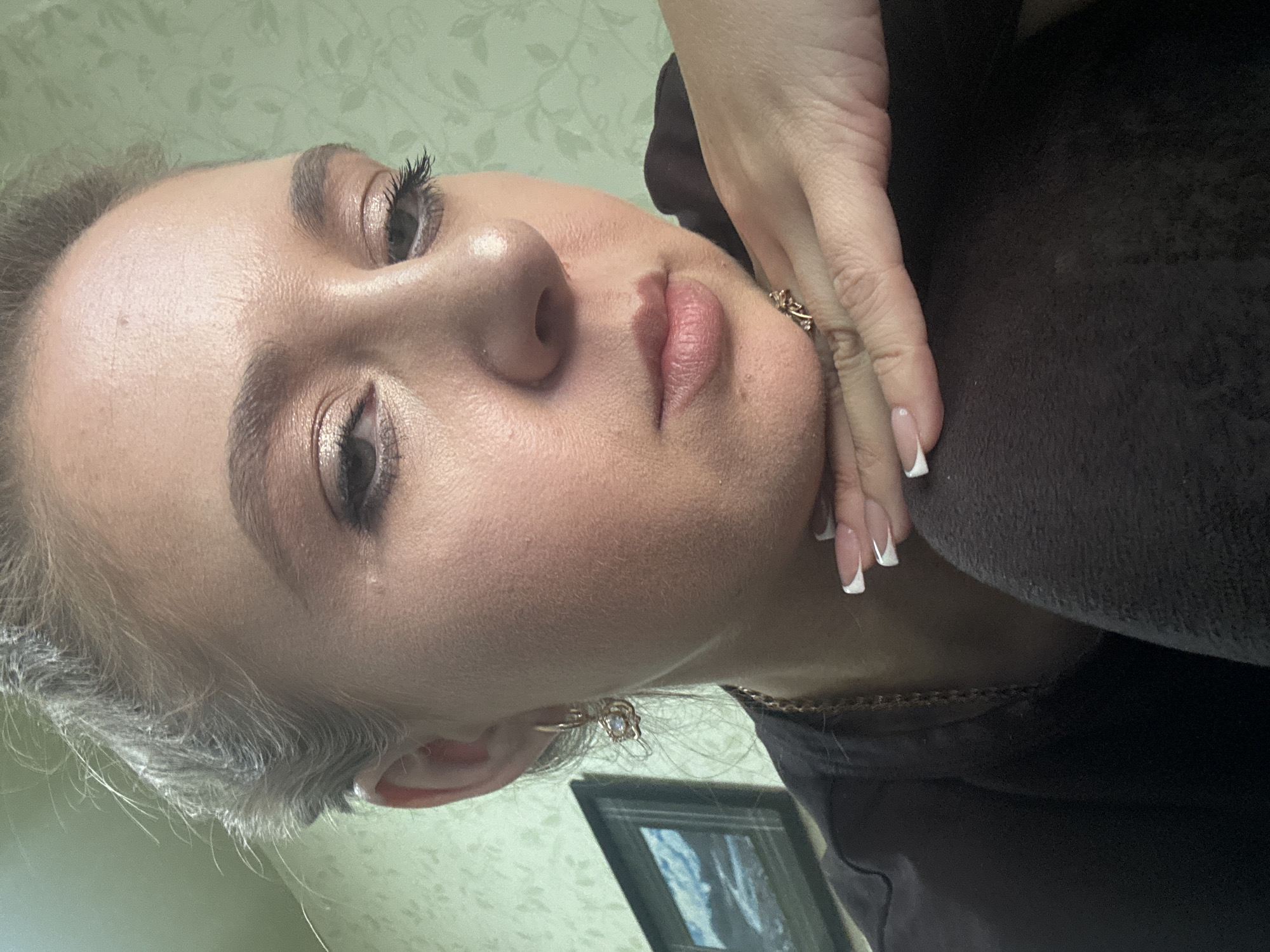 A close-up portrait of a woman's face is captured from a slightly high angle, with her head tilted downwards and her chin resting on her hand. Her light brown hair is pulled back, with wisps framing her face. She wears subtle makeup, including eyeshadow and mascara, and a faint blush on her cheeks. Her lips are slightly parted, revealing a hint of pink lipstick. A delicate gold earring with a small gem dangles from her left ear. She is wearing a dark-colored shirt and a gold chain necklace. Her right hand, adorned with a ring, supports her chin, and her fingernails are painted with a French manicure. The background is a light green wall with a subtle floral pattern. A framed painting of a landscape is visible on the wall to her left. The lighting is soft, highlighting the contours of her face and casting gentle shadows.
