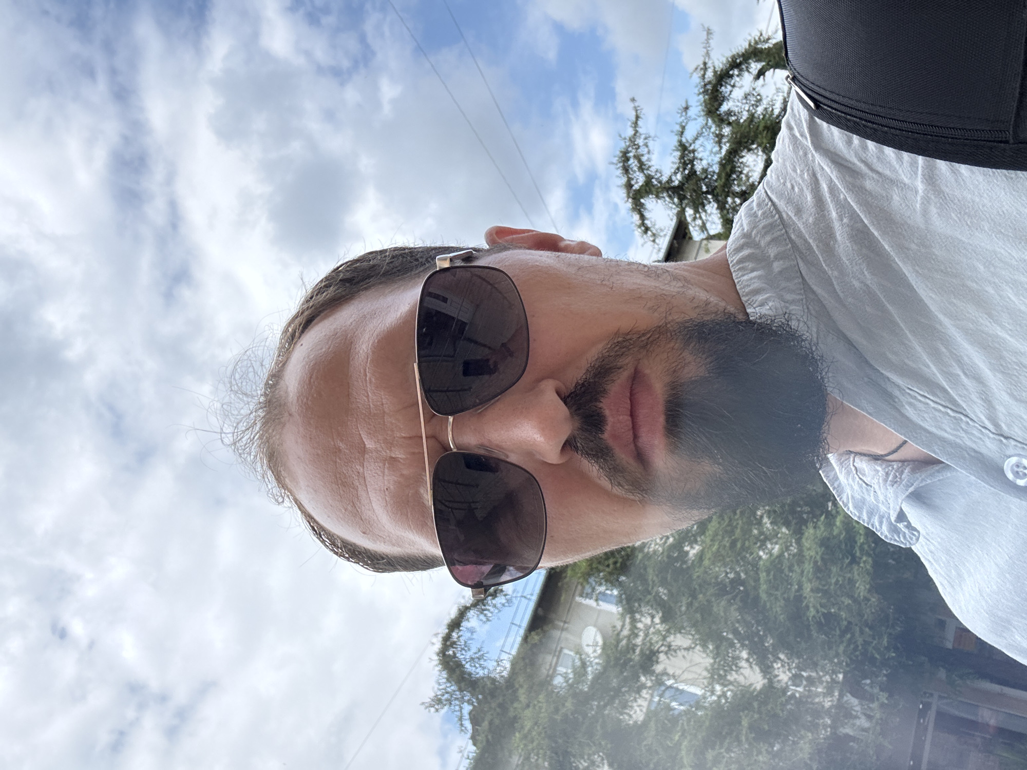 This is a close-up, low-angle selfie taken outdoors. The main subject is a man with a beard, wearing sunglasses and a light-colored collared shirt. He is looking towards the camera, and his face takes up a significant portion of the frame. The background consists of a cloudy sky with visible power lines and the branches of a tree. A black backpack strap is also visible over his shoulder. The lighting suggests it is daytime, possibly overcast due to the clouds. The image appears to be taken in an urban or suburban setting, given the presence of buildings and power lines in the blurred background.