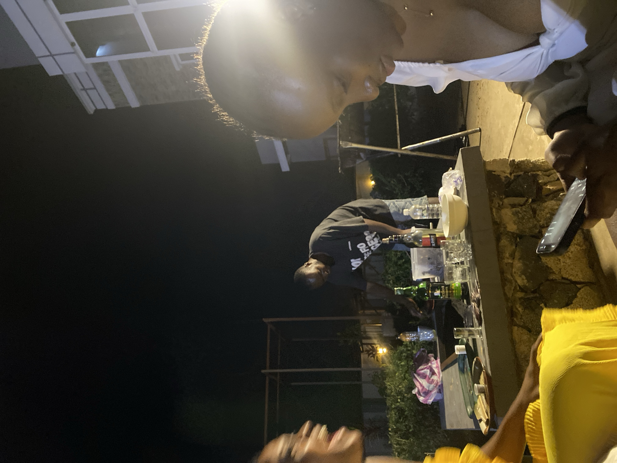 The image depicts a nighttime scene outdoors, possibly at a residential property in Gura Top, Nigeria. The primary focus is on a group of people gathered around a stone counter or table. In the foreground, on the right, a woman wearing a white top is looking down at a mobile phone. Her head is tilted, and her hair is short. To her left, a woman in a bright yellow outfit is also visible, her face blurred with motion and her mouth open as if speaking or laughing. In the background, a man wearing a dark t-shirt with white text is standing and appears to be preparing drinks or food. He is leaning over the counter.
On the stone surface, there are several objects, including bottles of alcohol, glasses, a bowl, and a plastic container. A green bottle labeled "Action Bitters" is prominent. A pink and white patterned cloth is also present. The background shows a dark sky and hints of a building with illuminated windows, suggesting an urban or suburban environment. The overall lighting is dim, with artificial lights contributing to the illumination of the scene. Lush green foliage is visible behind the counter, indicating a garden or outdoor seating area.