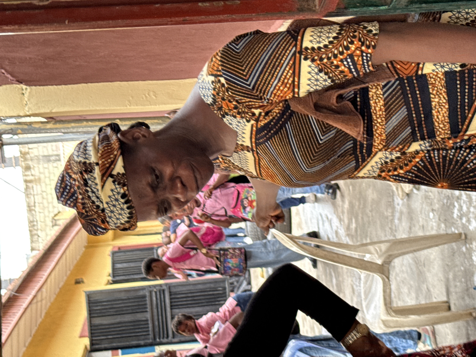 In Ado, Nigeria, an eye-level shot depicts a brown-skinned woman wearing a patterned headscarf and a yellow and brown patterned dress. She is bent over, smiling. In the background, several people wear pink shirts, many with blue jeans. There is a white plastic chair in front of the woman. A partially visible building with a yellow exterior is on the left. Above her, a red ceiling overhangs.