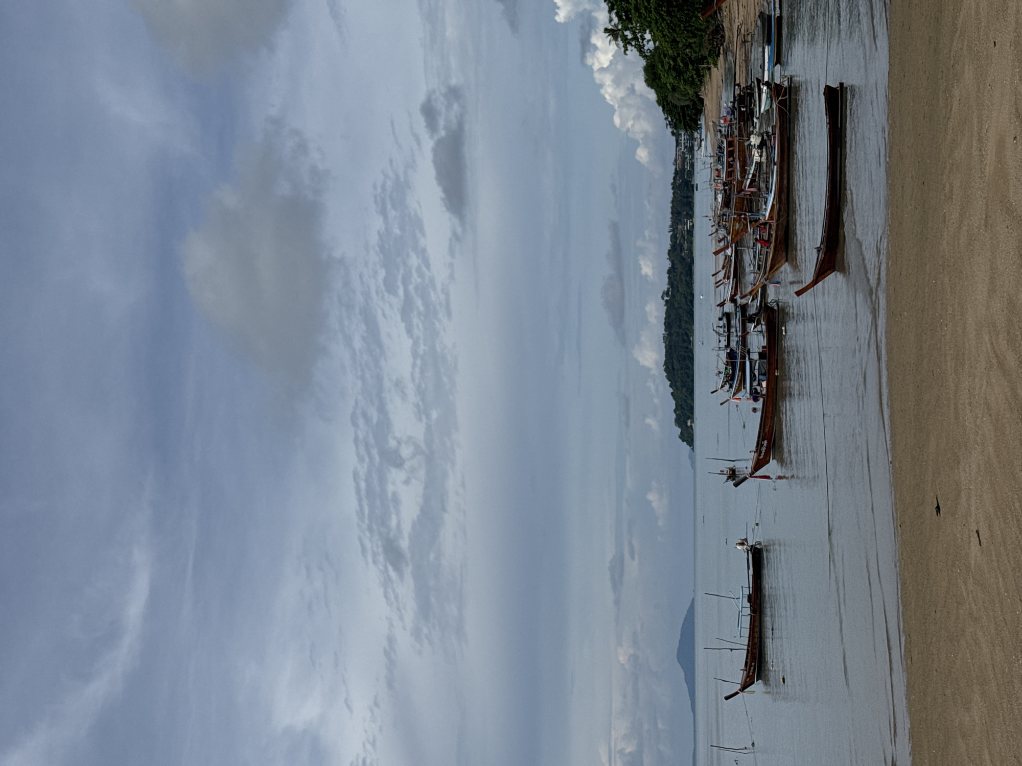 This is a vertical, full shot of a beach with a cloudy sky. The foreground is a sandy beach, with a few small pieces of debris. The middle ground is the calm, gray ocean, filled with numerous long, narrow wooden boats with pointed prows, many tied together or to poles. In the background, a densely vegetated coastline with buildings is visible, and further in the distance, a hazy, mountainous island under a sky filled with variegated clouds.