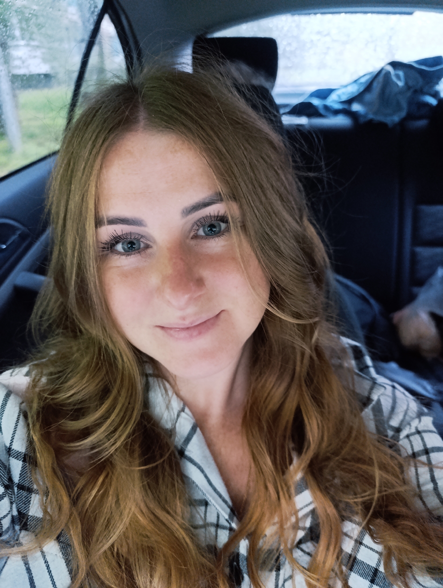 A woman with long, wavy blonde hair and blue eyes is taking a selfie inside a vehicle. She is wearing a plaid shirt. The background suggests she is in the back seat of a car, with the window to her left showing a blurred view of trees and a cloudy sky. The interior of the car is dark, with dark upholstery and a hint of blue fabric in the background. The image appears to have been taken in Chernogolovka, Russia.