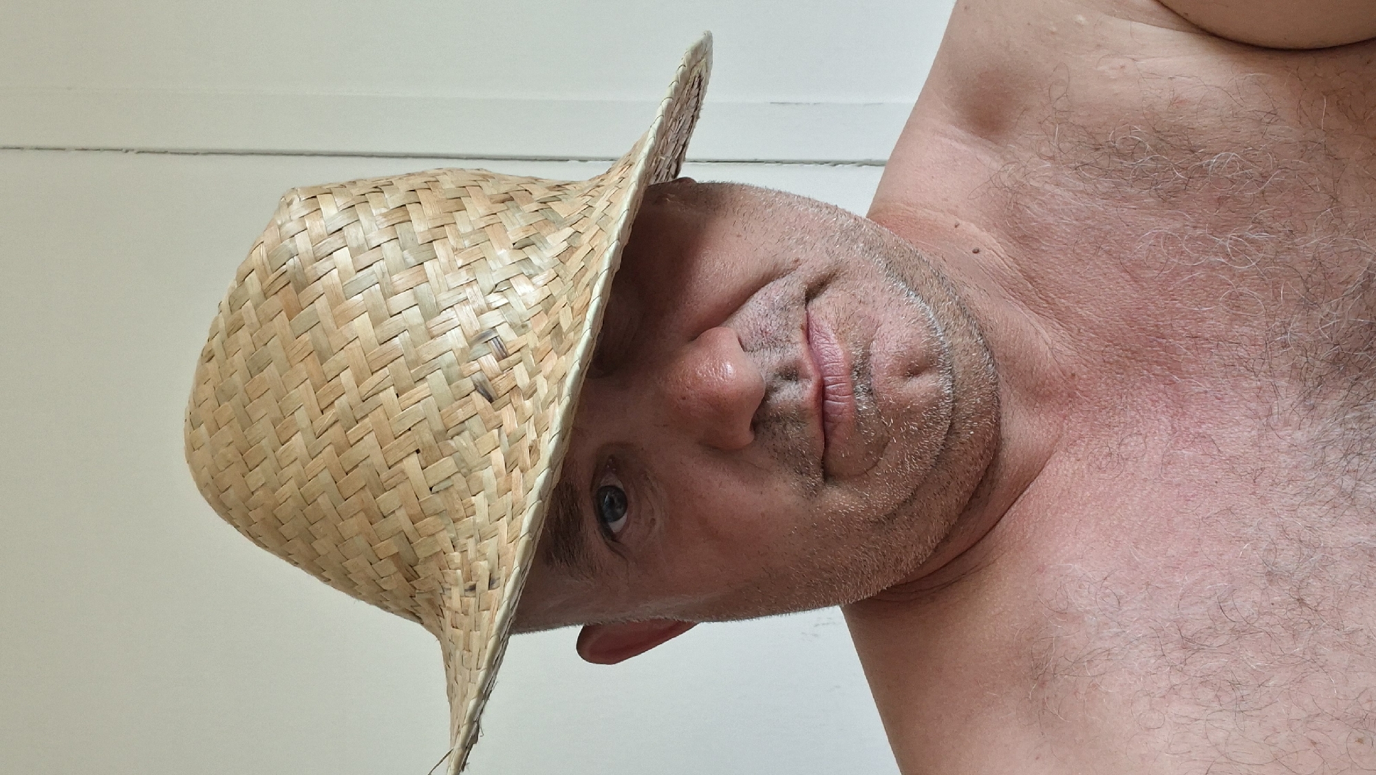 The media file is a close-up, low-angle, rotated photograph of a man wearing a straw hat. The man's face is partially obscured by the hat, with only his eye, nose, and mouth visible. He has stubble and appears to be looking towards the camera. His bare chest and shoulder are visible, with some chest hair and skin detail. The background is a plain, light-colored wall. The lighting suggests it could be daytime. The overall impression is a casual, possibly humorous, self-portrait. No text is visible in the image. The location context provided is Hîncești, Moldova.