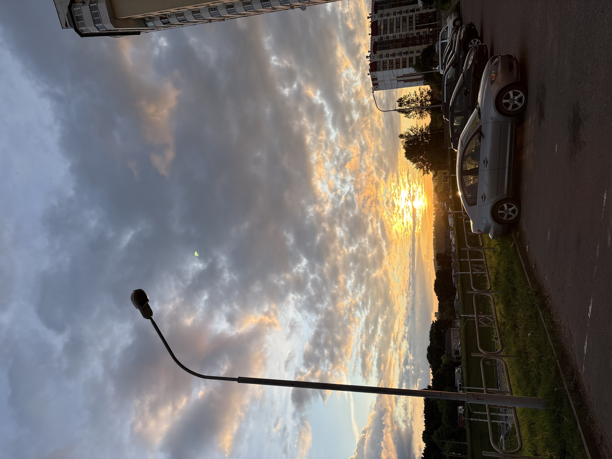 The image depicts a sunset over a parking lot in Vitsebsk, Belarus. A row of cars, including a silver sedan in the foreground, are parked next to a sidewalk and grassy area. A streetlamp stands to the left of the frame, casting a shadow. In the background, a tall residential building and a shorter building with red accents are visible, along with a tree line. The sky is filled with dramatic clouds illuminated by the setting sun, creating a warm, orange and yellow glow. The overall scene is one of an urban environment at dusk, with a natural spectacle in the sky.
