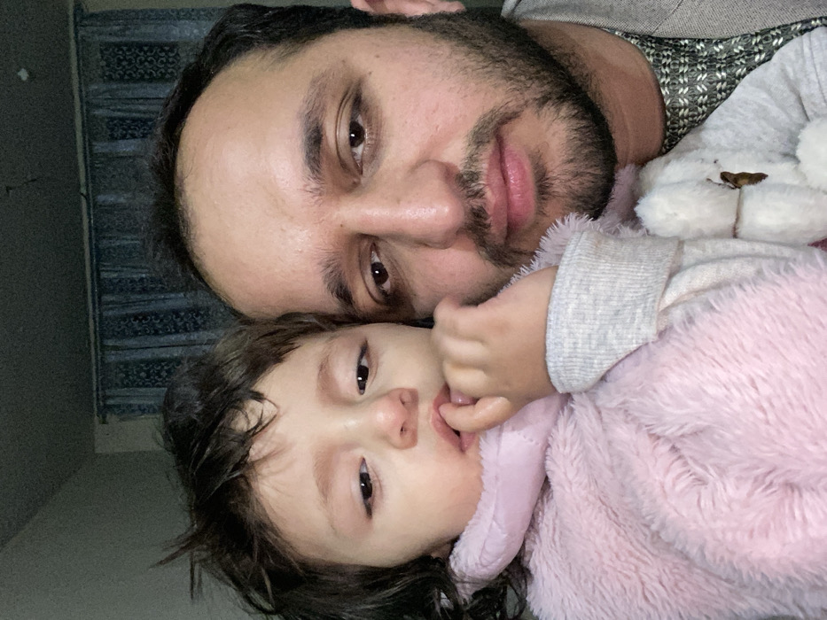 A man with a beard and dark hair is pictured in a close-up shot, lying next to a young child. The child, wearing a fuzzy pink jacket, has dark hair and brown eyes, and appears to be sucking on their fingers. A stuffed bear is also visible, nestled beside the man. The photo is taken at a slightly angled perspective.