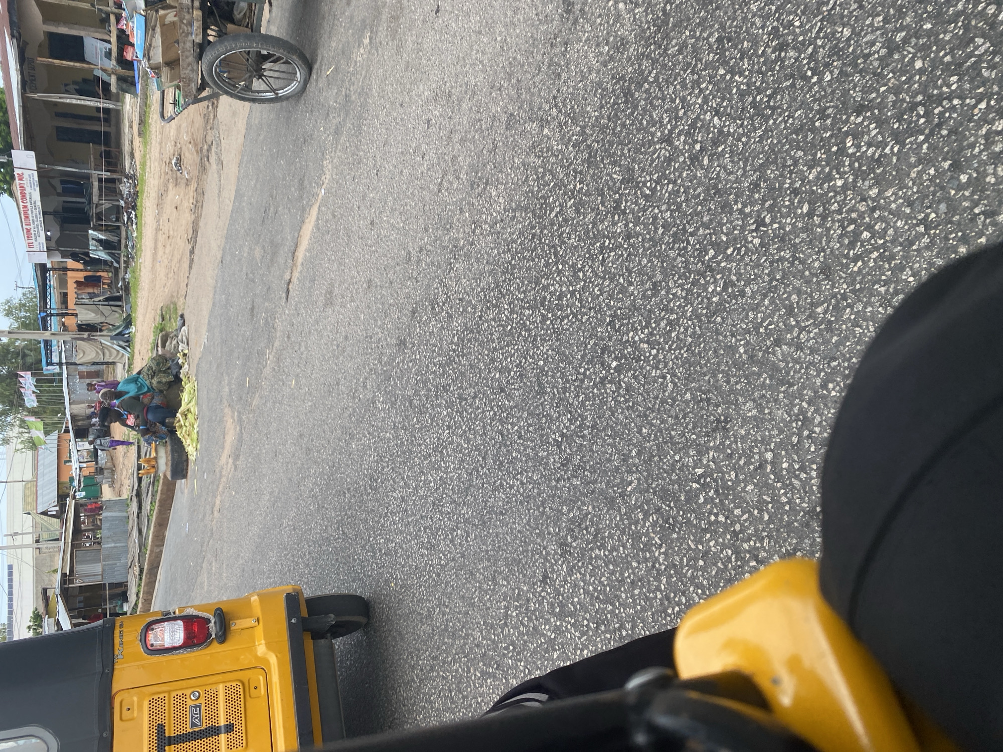 This image depicts a street scene in Jalingo, Nigeria. The foreground features a yellow three-wheeled vehicle, likely a tuk-tuk, with a black canopy. Part of the driver's black attire is visible in the lower right corner, as is a portion of the vehicle's yellow frame. The vehicle is positioned on a grey asphalt road.
To the left, the scene transitions to a roadside market area. Several people are gathered, some of whom appear to be vendors or customers. They are standing near a pile of what looks like agricultural produce, possibly corn or stalks, and some fabric items are draped nearby. The buildings in this area are simple structures, with some appearing to be shops or stalls. One building has a sign that reads "ITI YOUNG ALUMINUM COMPANY INC. WE DEAL IN ALL GLASS & ALUMINUM DOORS, WINDOWS AND FRAMES. ALUMINUM FABRICATION." Faded banners or flags hang from some of the structures.
The ground in the market area is dirt. The overall impression is of a bustling, everyday scene in a Nigerian town. The lighting suggests it could be daytime, possibly late morning or afternoon, with bright sunlight casting shadows. The weather appears to be clear.