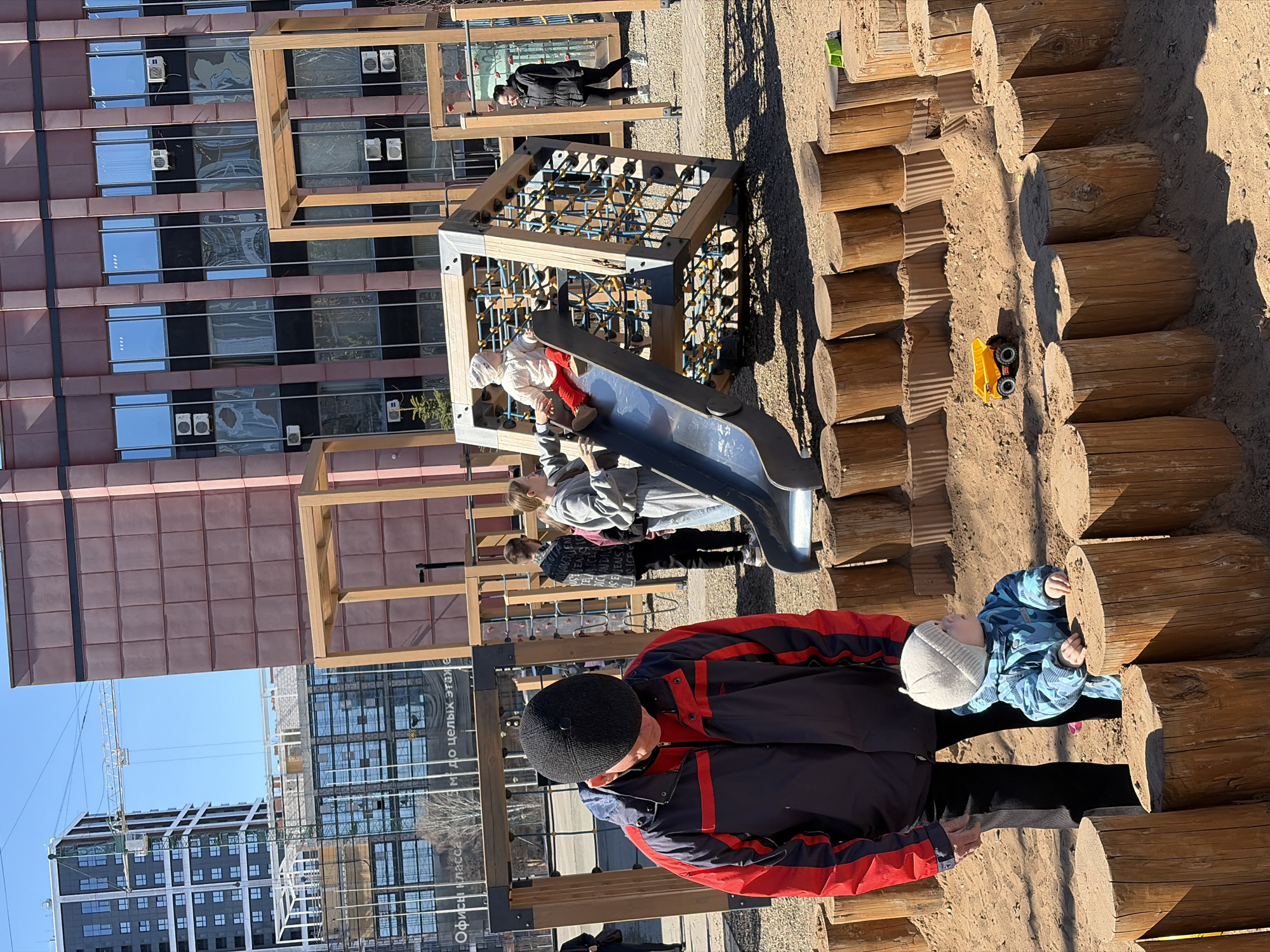 The image shows an outdoor playground scene in Izhevsk, Russia. A man wearing a black hat and a black jacket with red accents is standing near a young child in a blue patterned jacket and grey hat, who is holding onto wooden logs. The logs form a path or obstacle course. Another group of children are near a slide and a climbing structure. In the background, there are buildings with reflective windows. A toy truck is visible on the logs.
