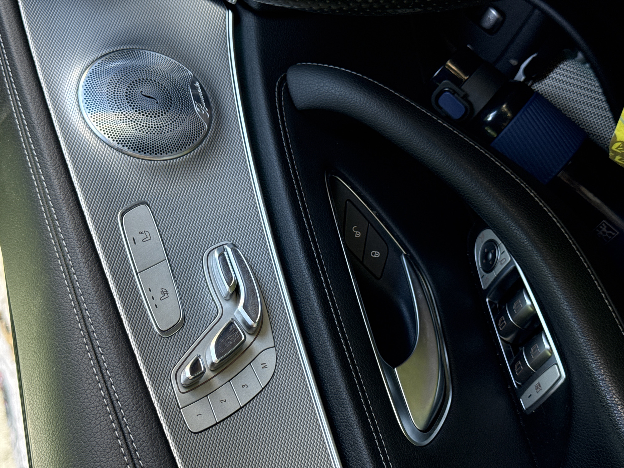 The image shows the interior of a car's driver's side door, featuring black leather upholstery with white stitching, silver trim panels, and a circular speaker grille with the "Burmester" logo. Control buttons for seat adjustment, window operation, and door locks are visible. A dark blue insulated cup with a blue lid is placed in a recessed compartment. The image does not provide any information about the city of Budva, Montenegro.