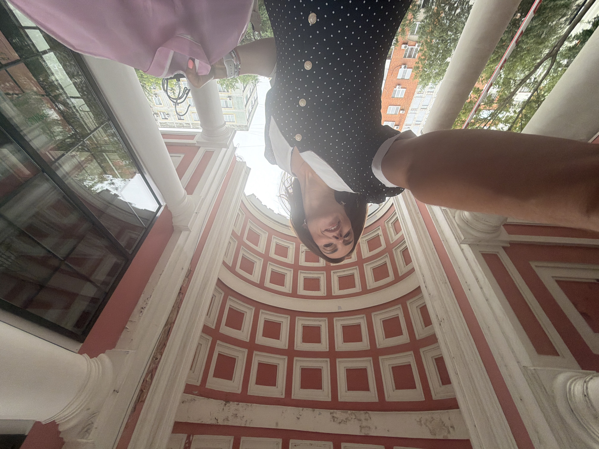 A woman is taking a selfie from a low angle in front of a building with a decorative red and white ceiling. She is wearing a black dress with white polka dots and a white collar. She is smiling and holding a pink garment on a hanger in her right hand. The background shows trees, other buildings, and a large window reflecting the sky and trees. The architectural style suggests a historical building, potentially a museum or a government building. It is daytime, and the weather appears to be overcast. The image was taken in the city of Khabarovsk, Russia.
