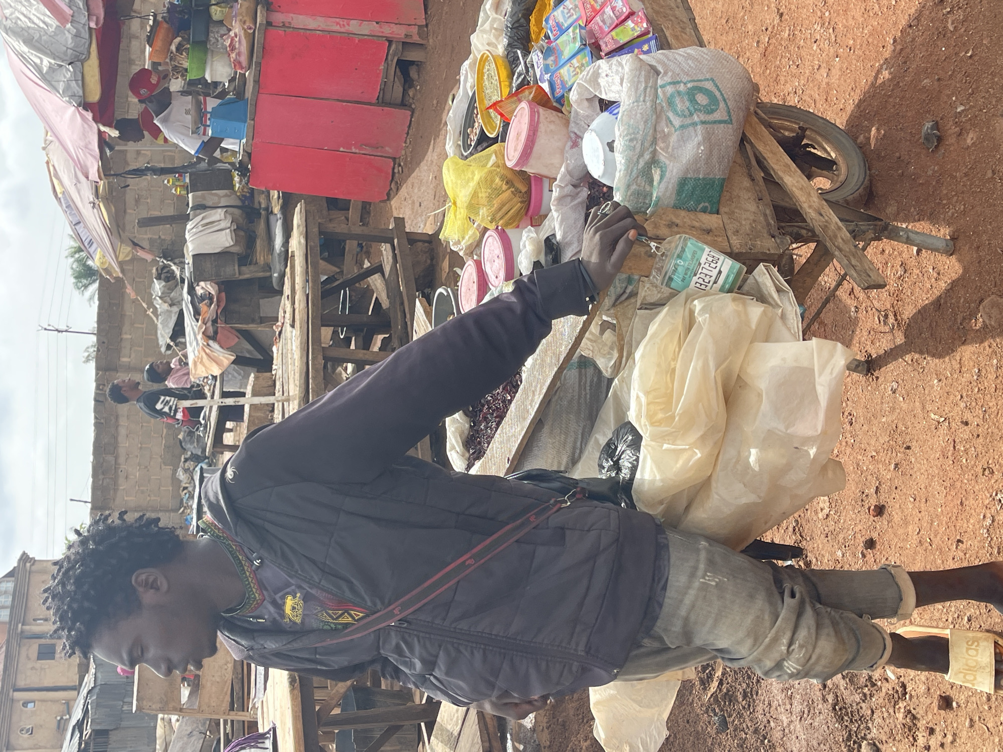 A man is captured in the midst of a marketplace setting, his presence suggesting a potential transaction or engagement with the goods at hand. He is dressed in a dark jacket, adorned with a colorful shirt, and paired with gray denim pants. His footwear consists of Adidas-branded sandals.
The scene is cluttered with a variety of items on a wooden cart, including bagged goods, pink containers, and packaged products. The environment points towards Gura Top, Nigeria, with the presence of red soil and distinct infrastructure. The context of the image appears to be of commerce, displaying the sale of goods in an outdoor setting.
