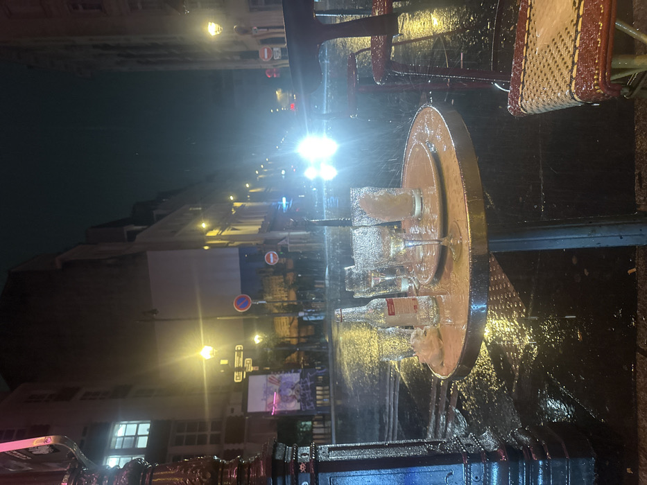 A street scene at night is soaked in rain. A small round table sits outside, covered in empty glasses and bottles. Rainwater streams off the table's edge. A red chair is partially visible. Streetlights illuminate the scene, creating a blurred, wet reflection on the pavement. Buildings line the street in the background, with various signs and lights visible. The overall atmosphere is one of a deserted, rainy night in a city.