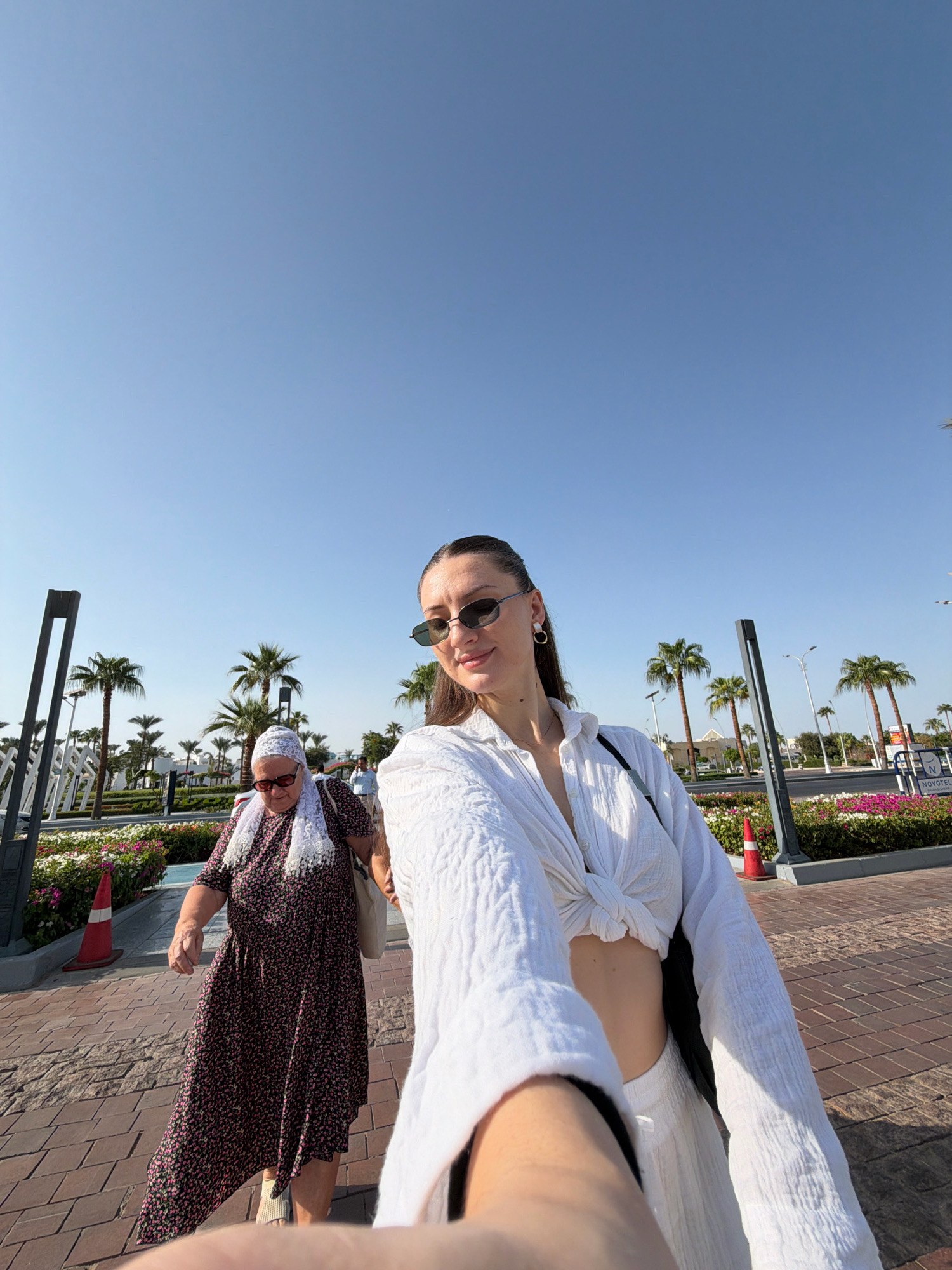 In Sharm El Sheikh, Egypt, an image captures a bright, sunny day with a clear blue sky. In the foreground, a young woman with brown hair pulled back is taking a selfie. She is wearing sunglasses, a white, textured, open-front shirt tied at the waist, exposing her midriff, and white pants. To her left, an older woman in a floral dress, headscarf, and glasses walks away from the camera, her body angled slightly towards the young woman. Palm trees line the background, with flowering plants and streetlights visible. The ground is paved with reddish-brown bricks.