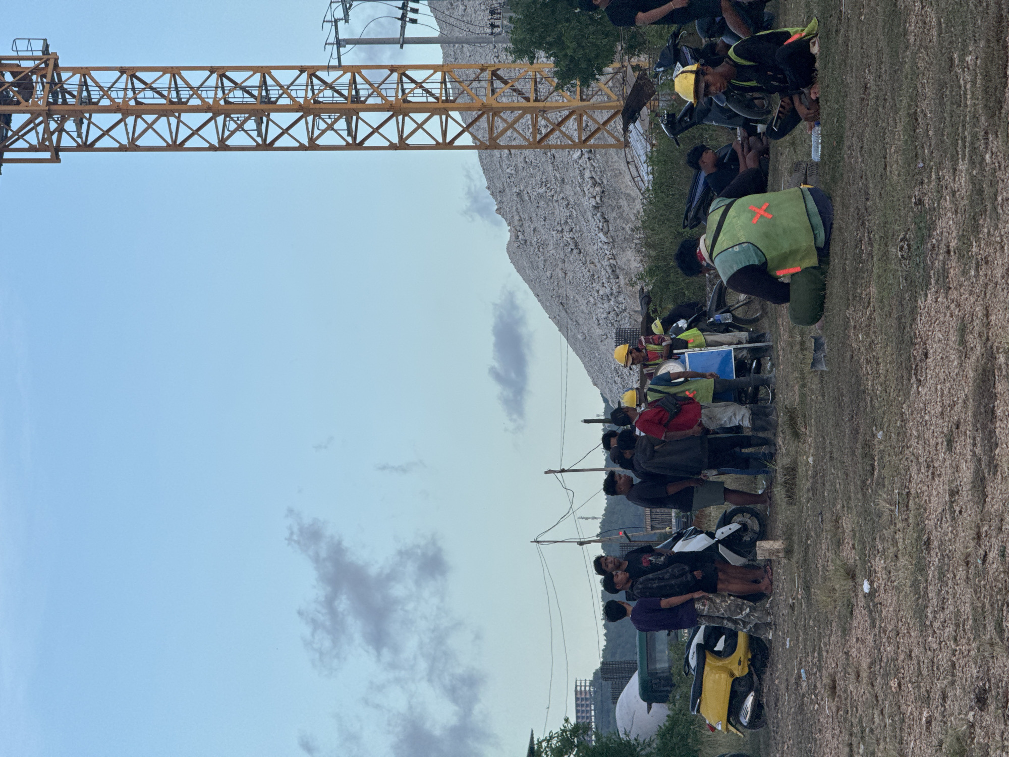 The image shows a group of people, mainly men, gathered on a grassy field. Many are wearing helmets and some have motorcycles parked nearby. A large yellow crane structure is visible in the background, along with what appears to be a rocky hillside or quarry. The sky is partly cloudy. The location is Kuta Selatan, Indonesia, based on the presence of people with motorcycles and the style of buildings. The group appears to be resting or taking a break, with some individuals sitting on the ground and others standing. Some are wearing reflective vests. There is trash around.