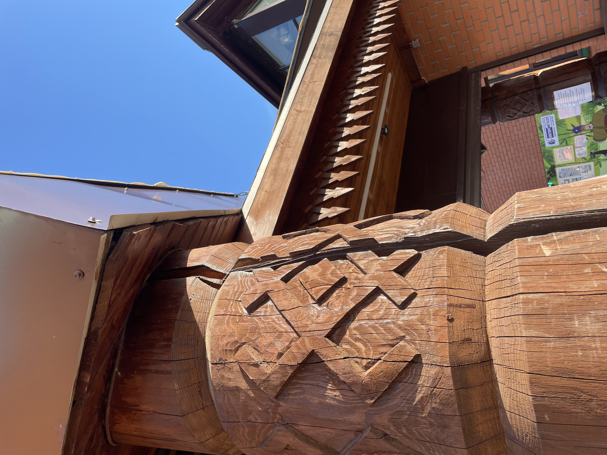 A low-angle outdoor shot depicts a segment of the Izhevsk Zoo's entrance in Izhevsk, Russia. A large, carved wooden beam with a zigzag pattern dominates the foreground, showing wood grain and aging cracks. To the left, a section of the roof with a transparent panel is visible against a clear blue sky. A wooden structure featuring a row of triangular decorations runs diagonally upwards. Further back, the brickwork of the building is seen alongside informational posters. The composition emphasizes architectural detail and the aged character of the wooden elements.